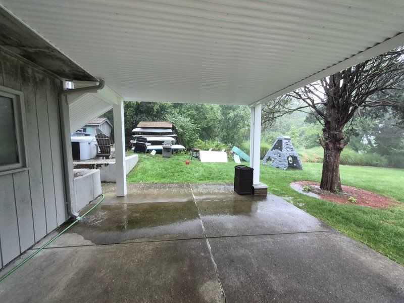 Covered concrete patio with wet floor, support post, lawn, trees, and a parked car in the yard.
