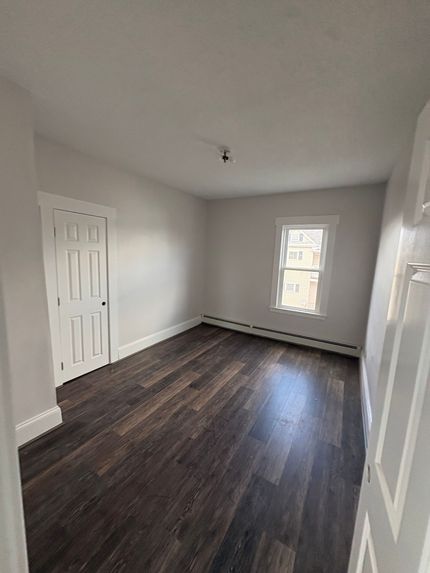 Empty white room with dark wood floors, a door on the left, and a window at the back.