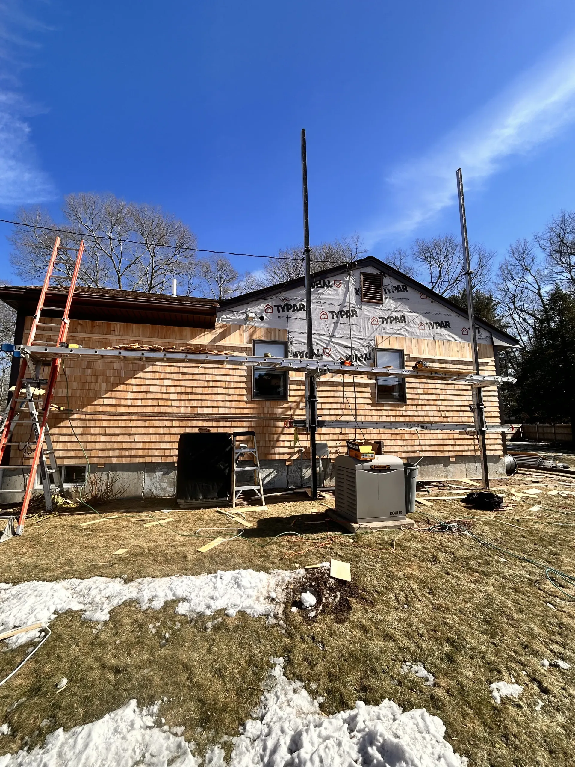 Partially clad wooden house under construction with ladders, chimneys, and patchy snow in the yard