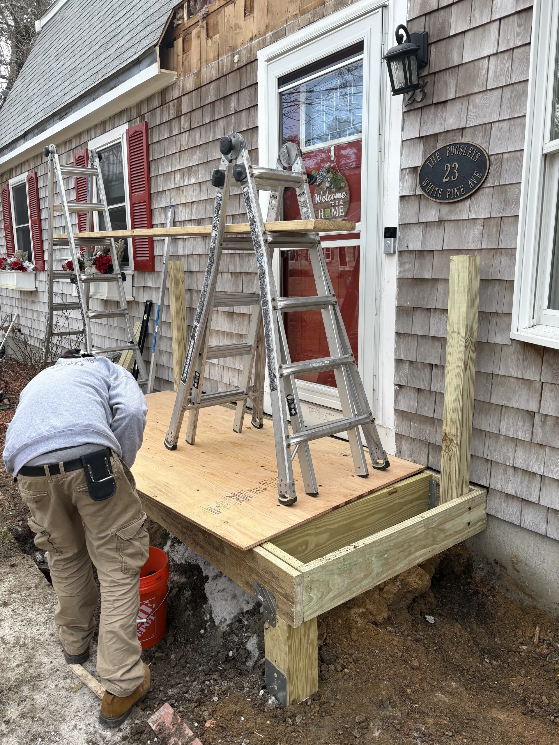 Worker on a small wooden platform beside a shingled house, handling ladders during exterior repair.