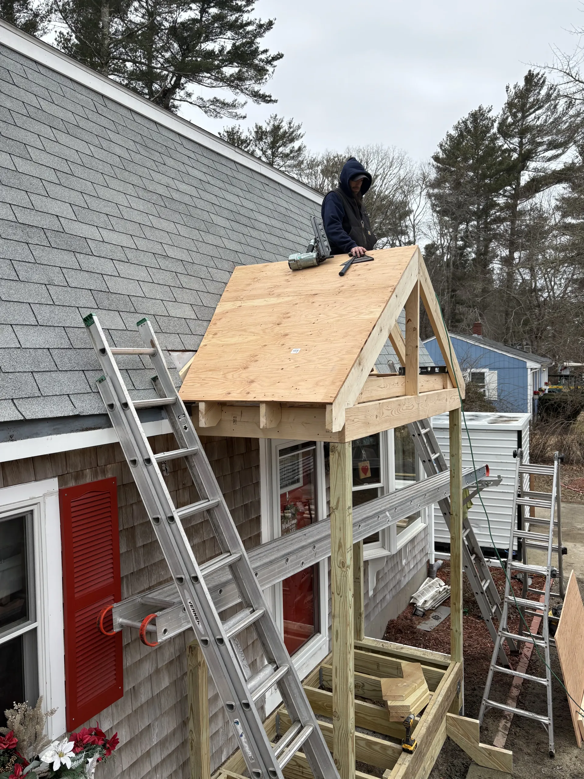 Worker on a ladder framing a small roof over a house entrance beside a brick wall