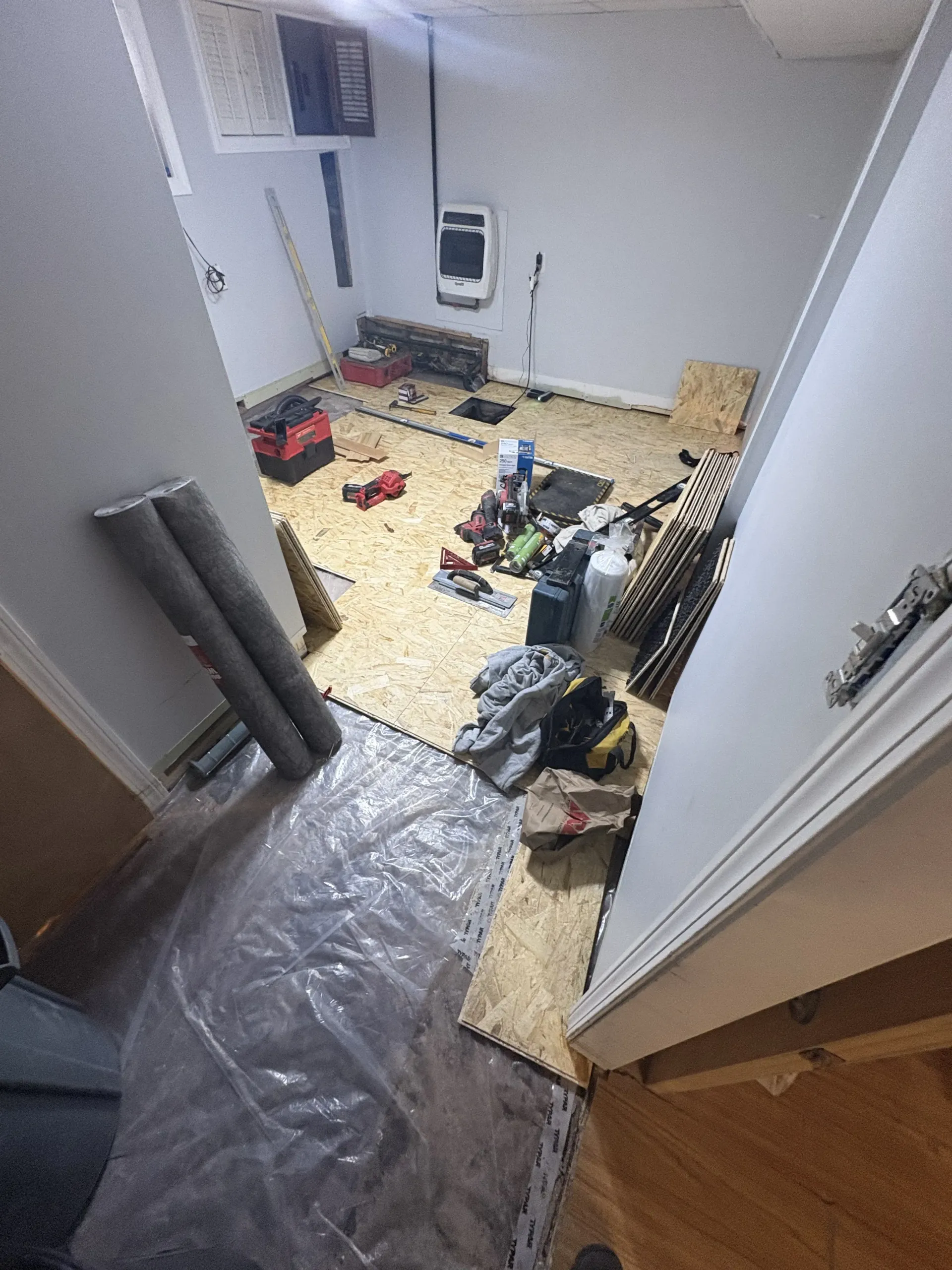 Cluttered room with tools, boxes, and plastic sheeting on the floor, viewed from a doorway