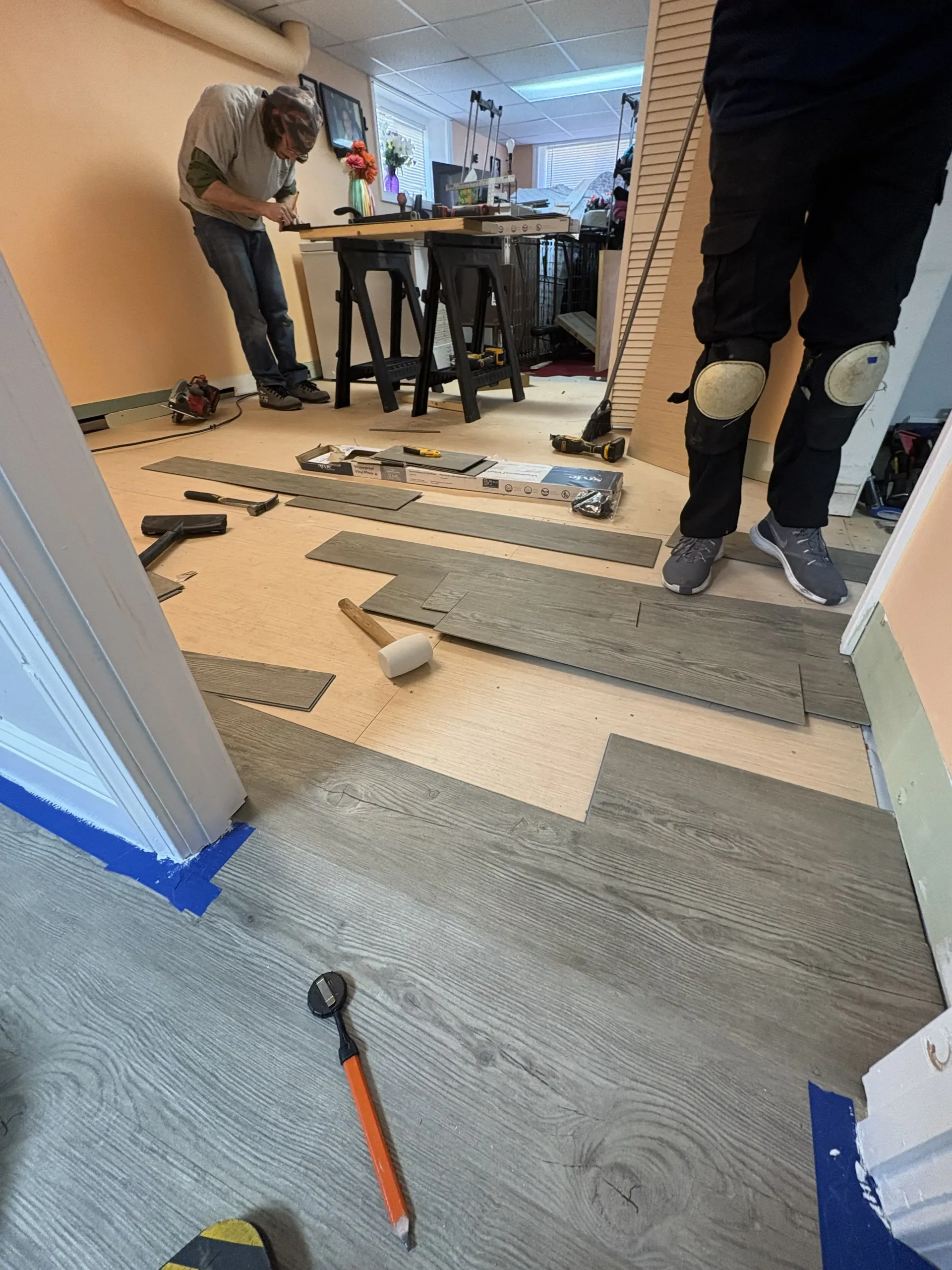 Workers installing flooring in a room, with tools and planks spread across the floor.