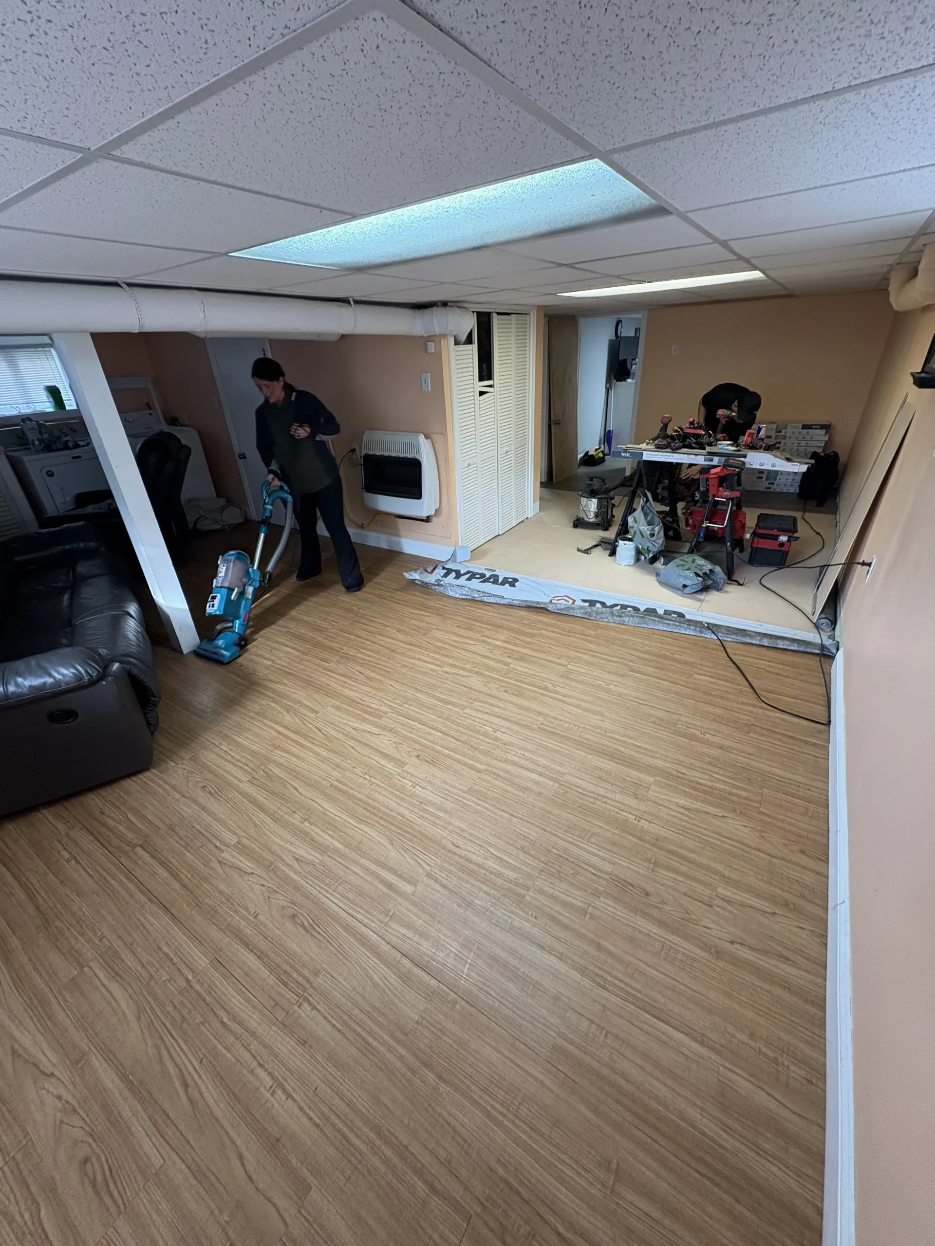 Basement room with wood floor, ceiling panels, and two people near a wall-mounted TV and exercise equipment