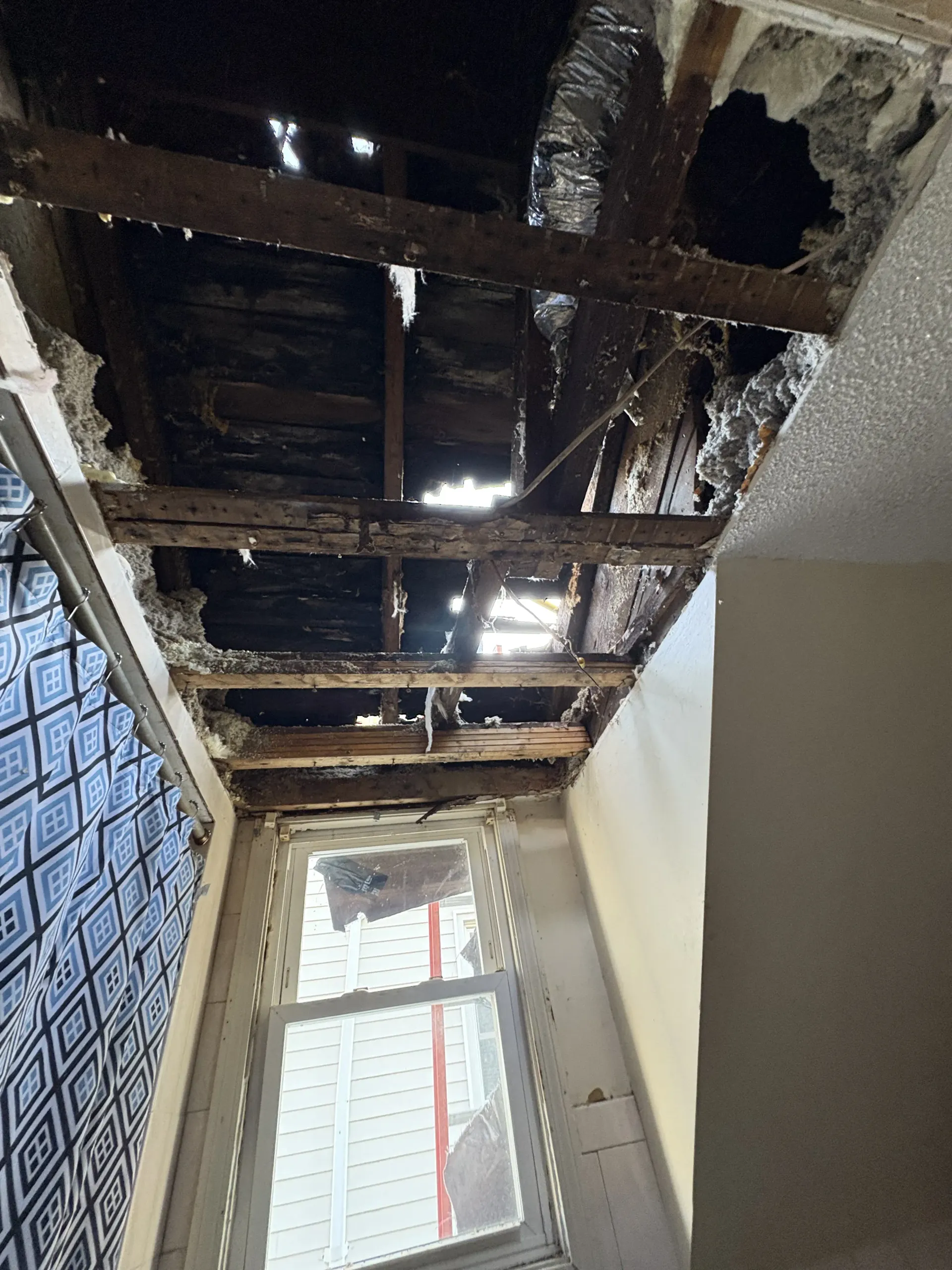Damaged ceiling with exposed wooden beams above a window in a narrow room