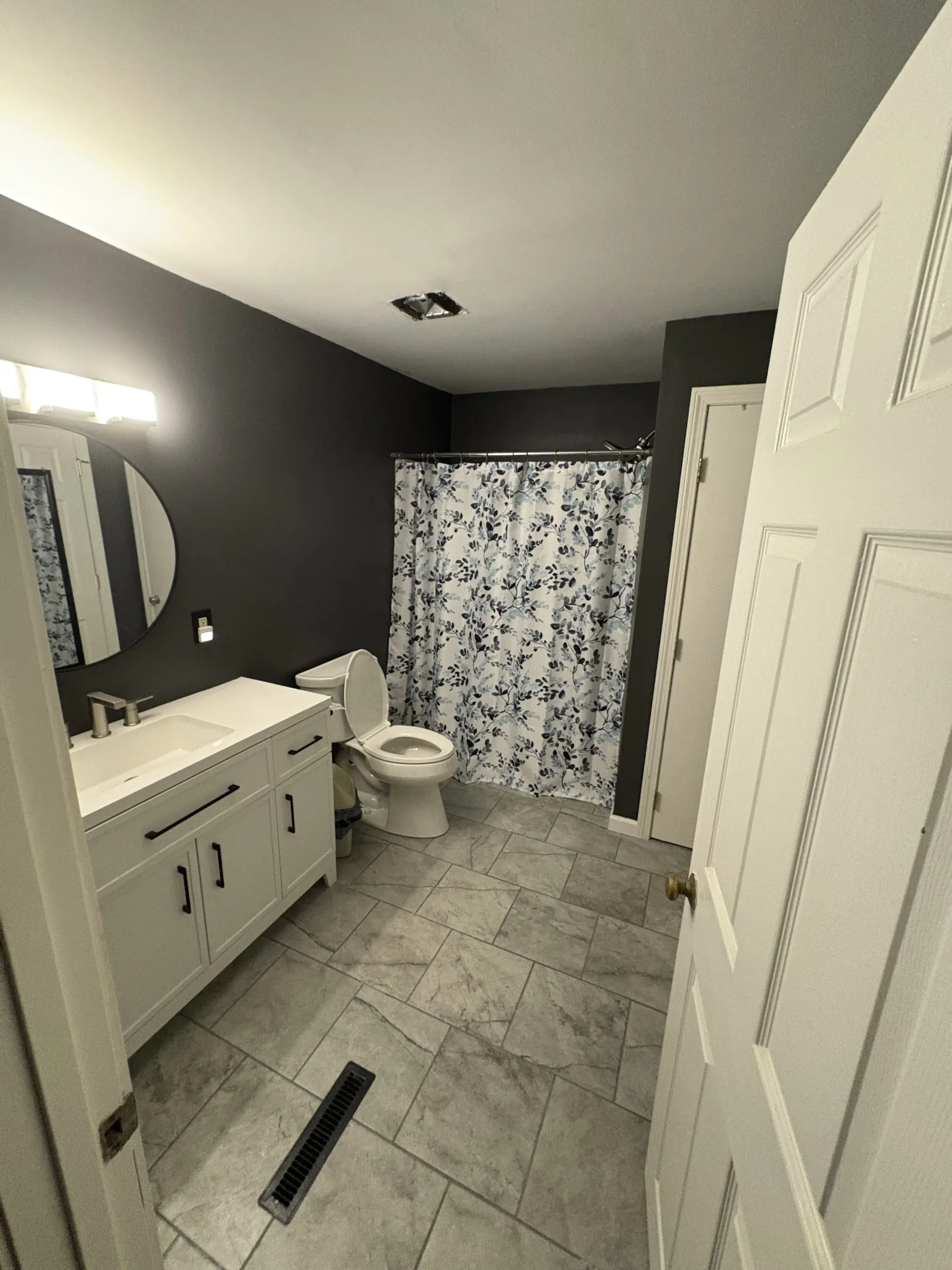 Small bathroom with gray tile, white vanity, toilet, and floral shower curtain under bright light.