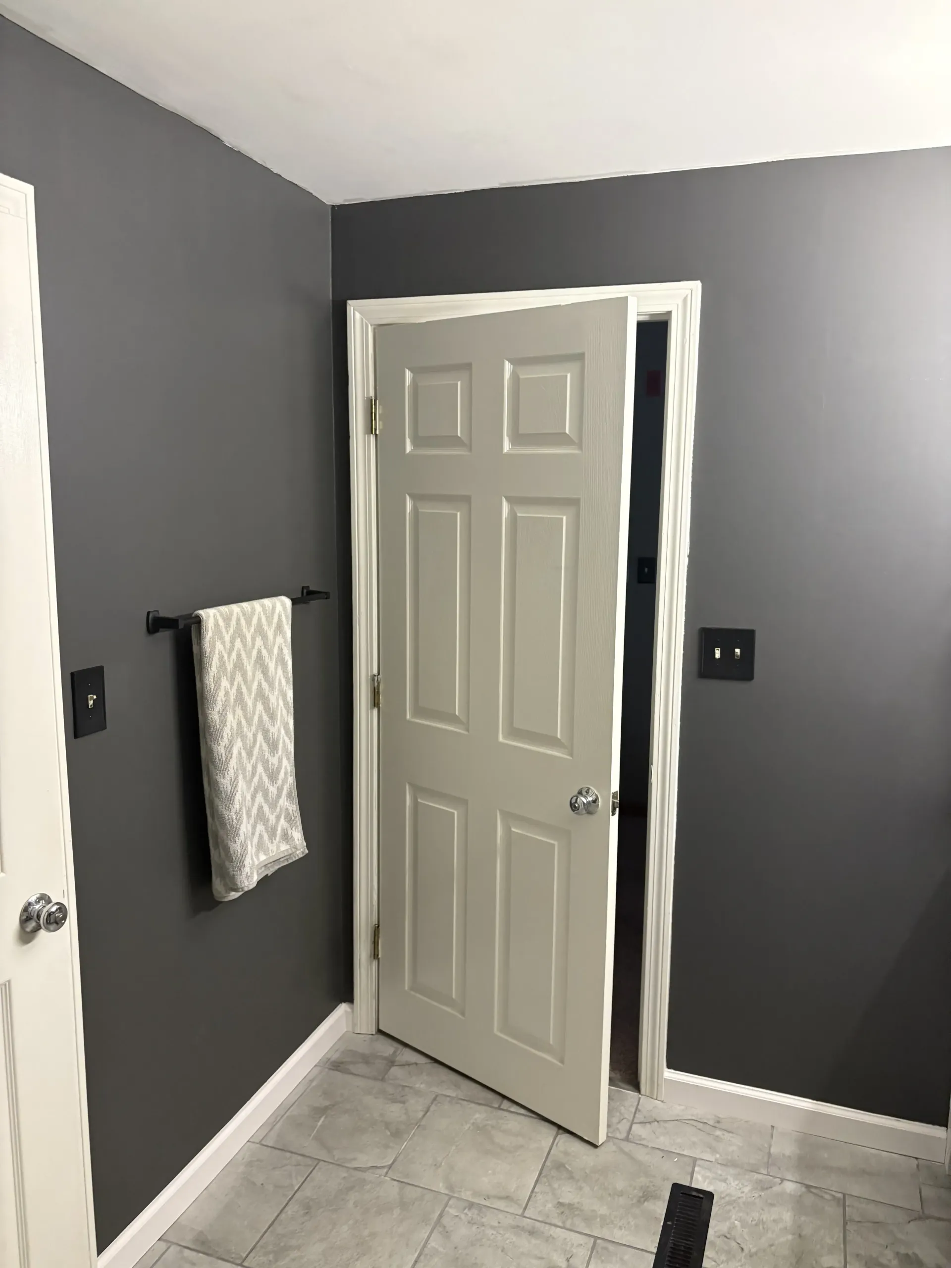 Gray bathroom with open white door, towel on wall, and tiled floor.