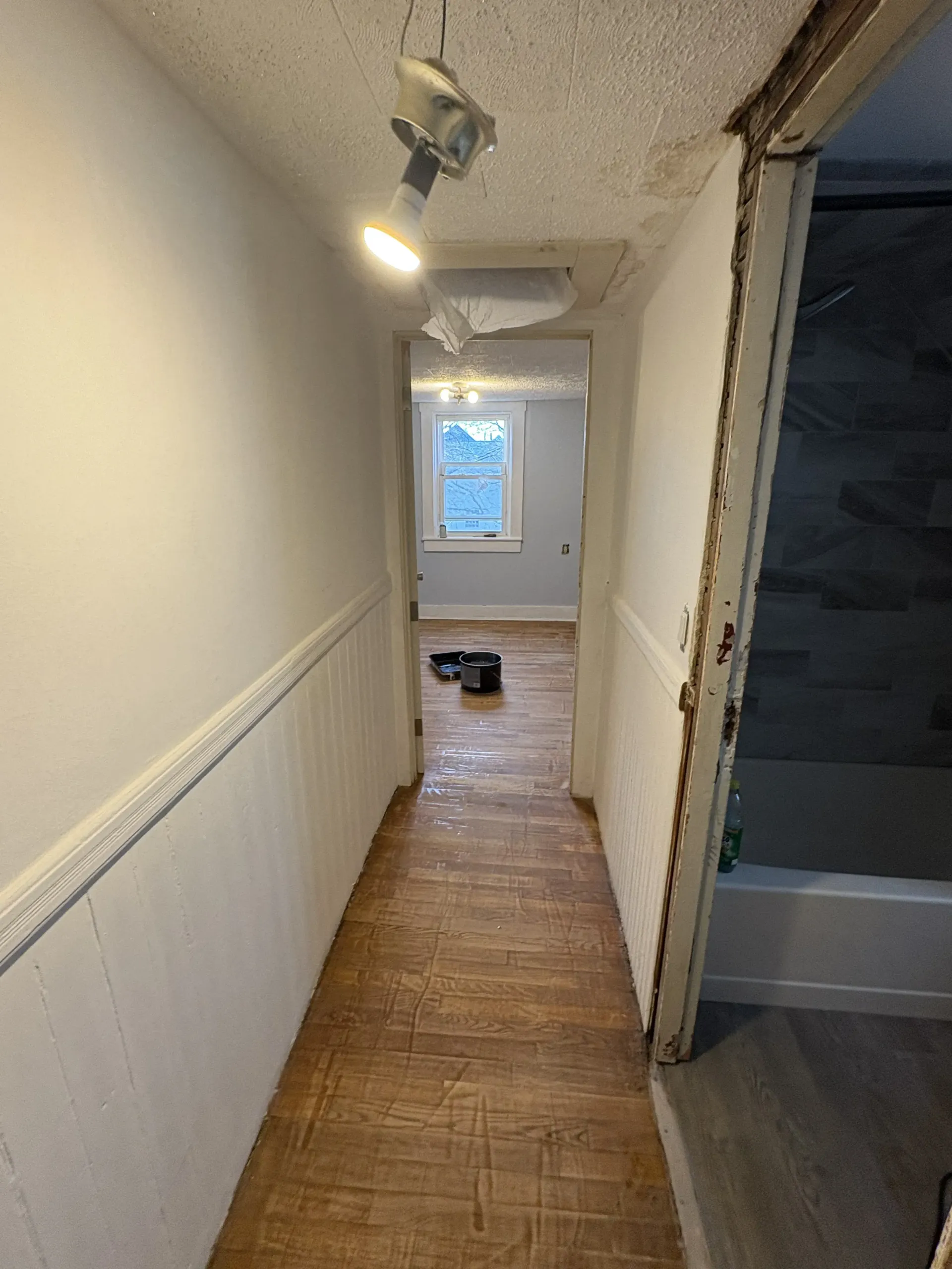 Narrow hallway with wood floor, white walls, and a small black item on the floor near a bright window.