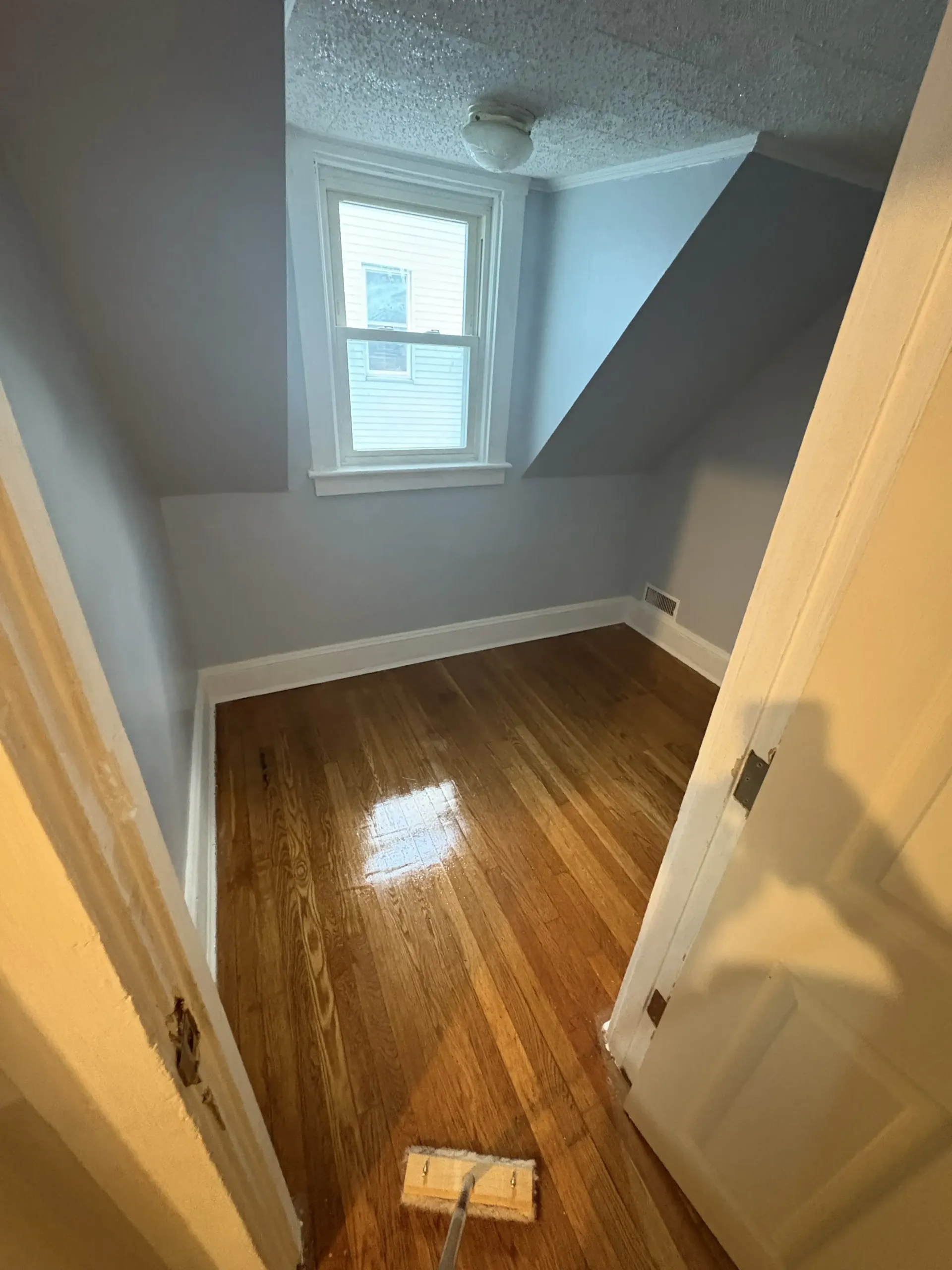 Small empty room with slanted ceiling, window, and glossy hardwood floor, viewed from the doorway.