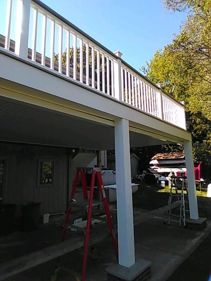 Covered deck with white railing and support posts over a shaded patio, with a red ladder below.