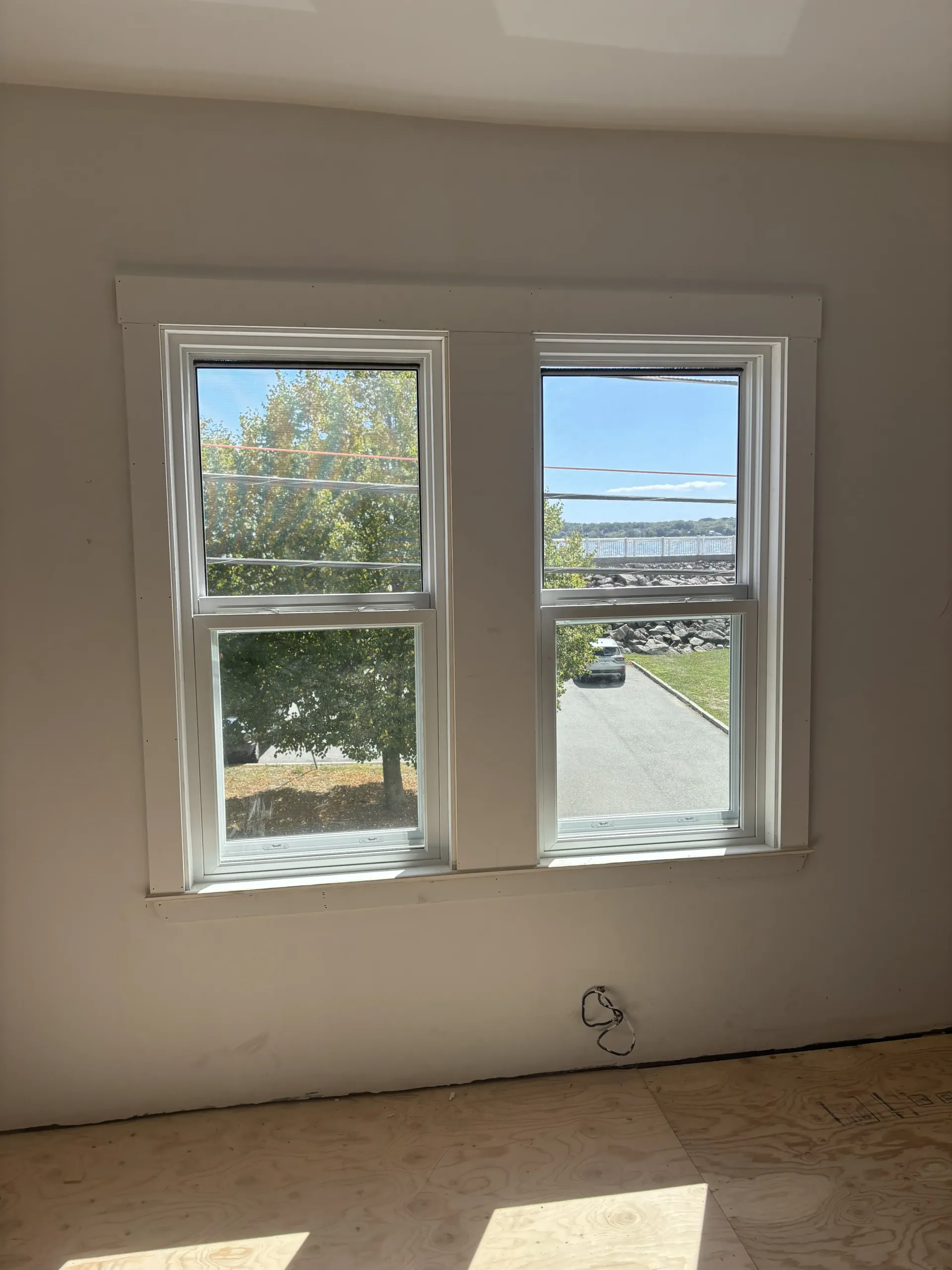 Empty room with white walls, two windows, and a view of trees, road, and water outside.