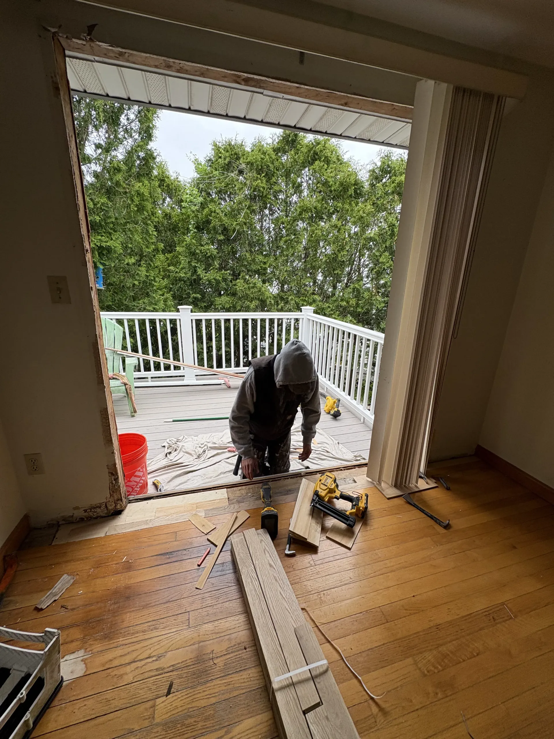 Person kneels on a wet porch outside an open doorway, with tools and a ladder inside a wood-floored room.