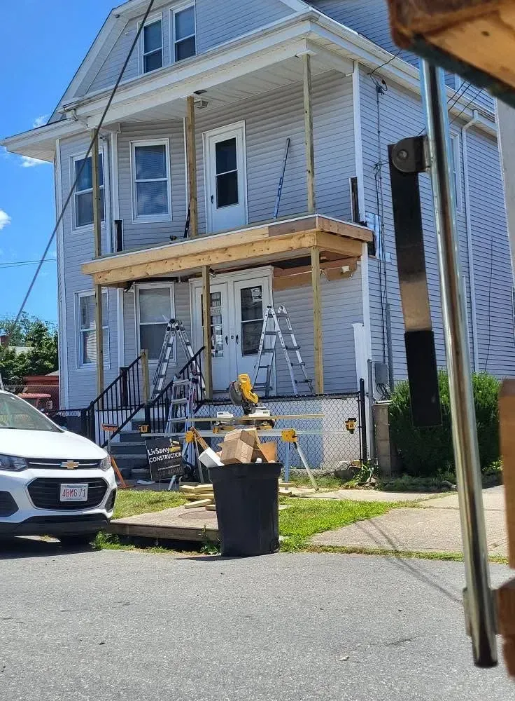 Two-story white house with front porch under construction, with ladders, wood, and a trash bin on the curb.