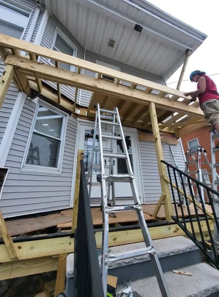 Workers building a wooden porch roof extension beside a house, with ladders and railings in place