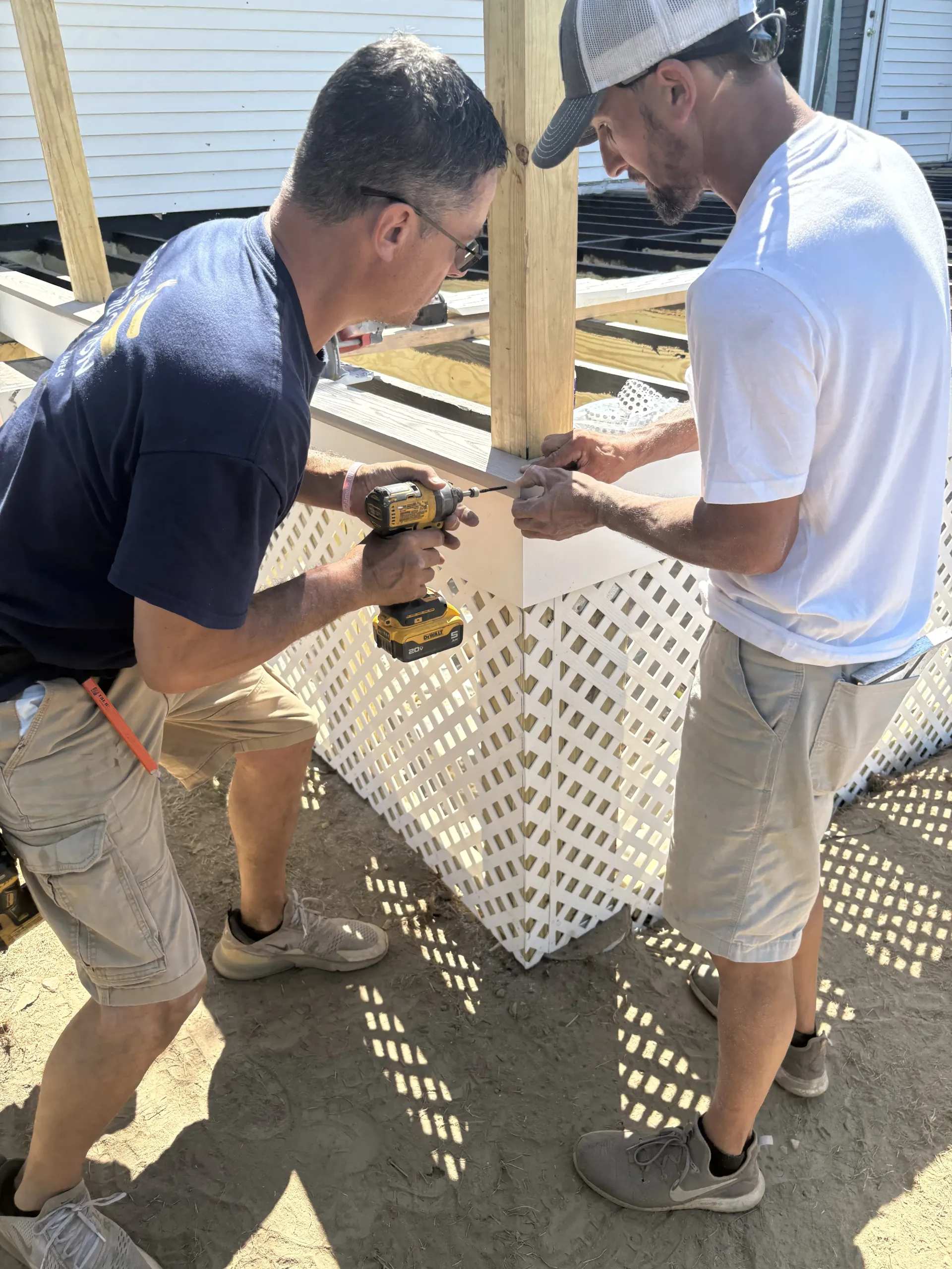 Two workers using power drills to assemble a white lattice structure outdoors.