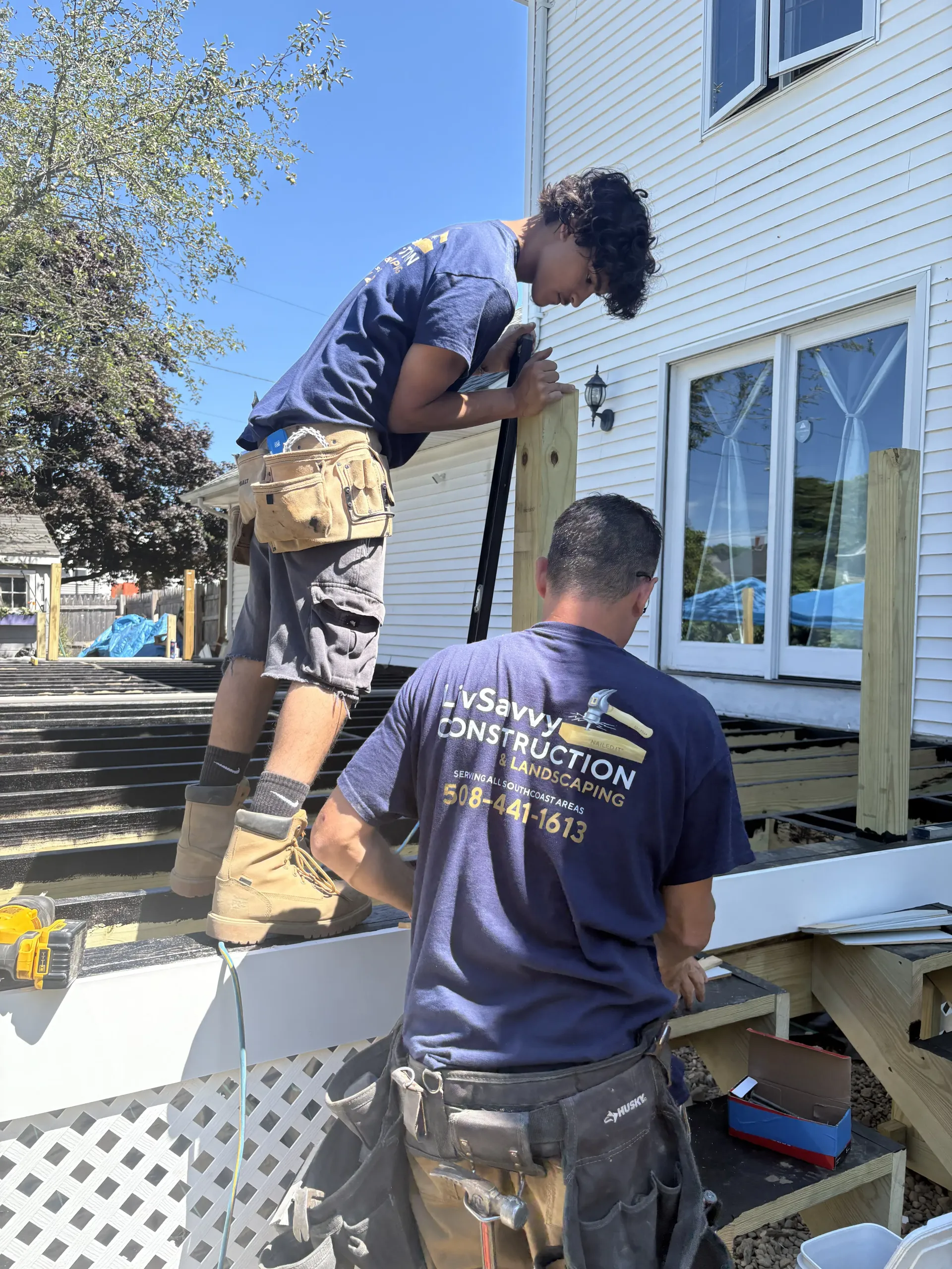 Two workers installing siding on a house exterior, one on a ladder beside a window.