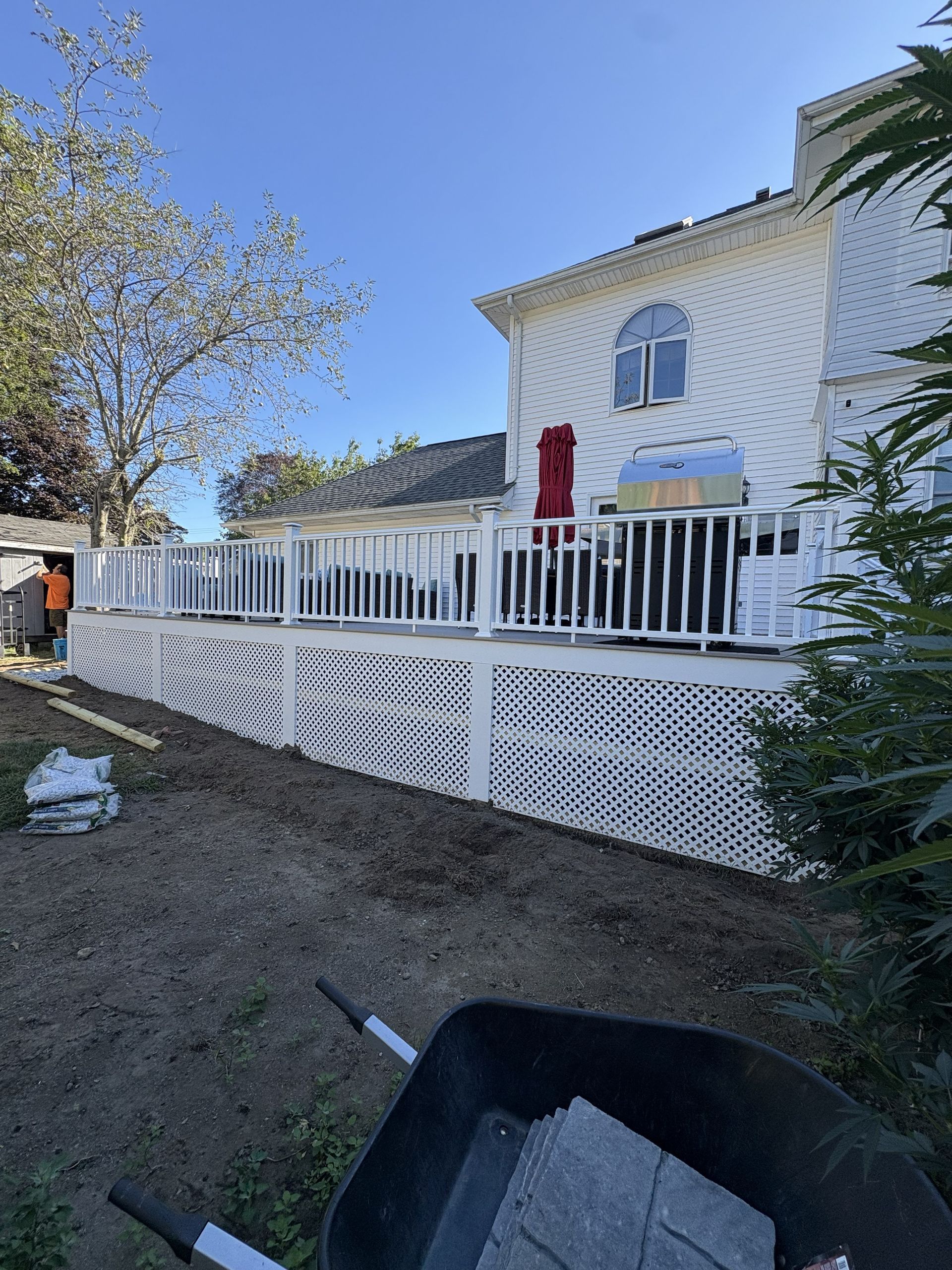 White house with a raised deck and black-and-white lattice railing in a yard with trees and gravel.