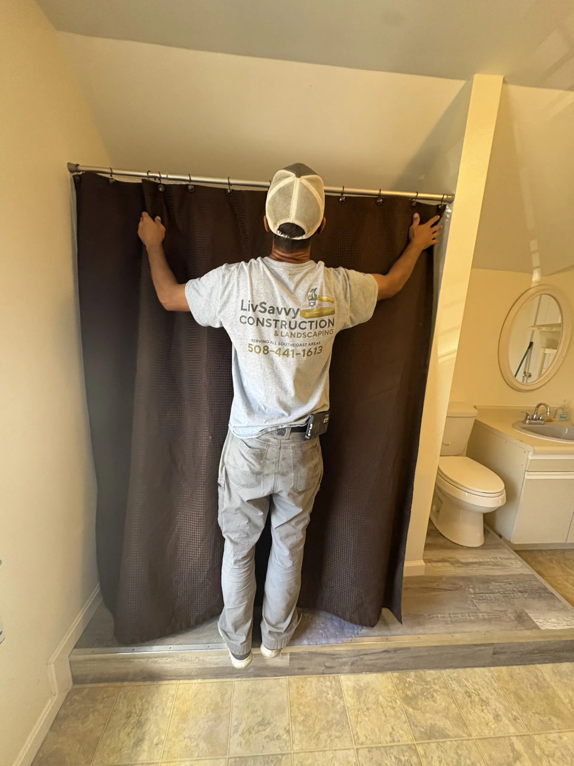 Person hanging dark shower curtains in a beige bathroom, beside a toilet and sink.
