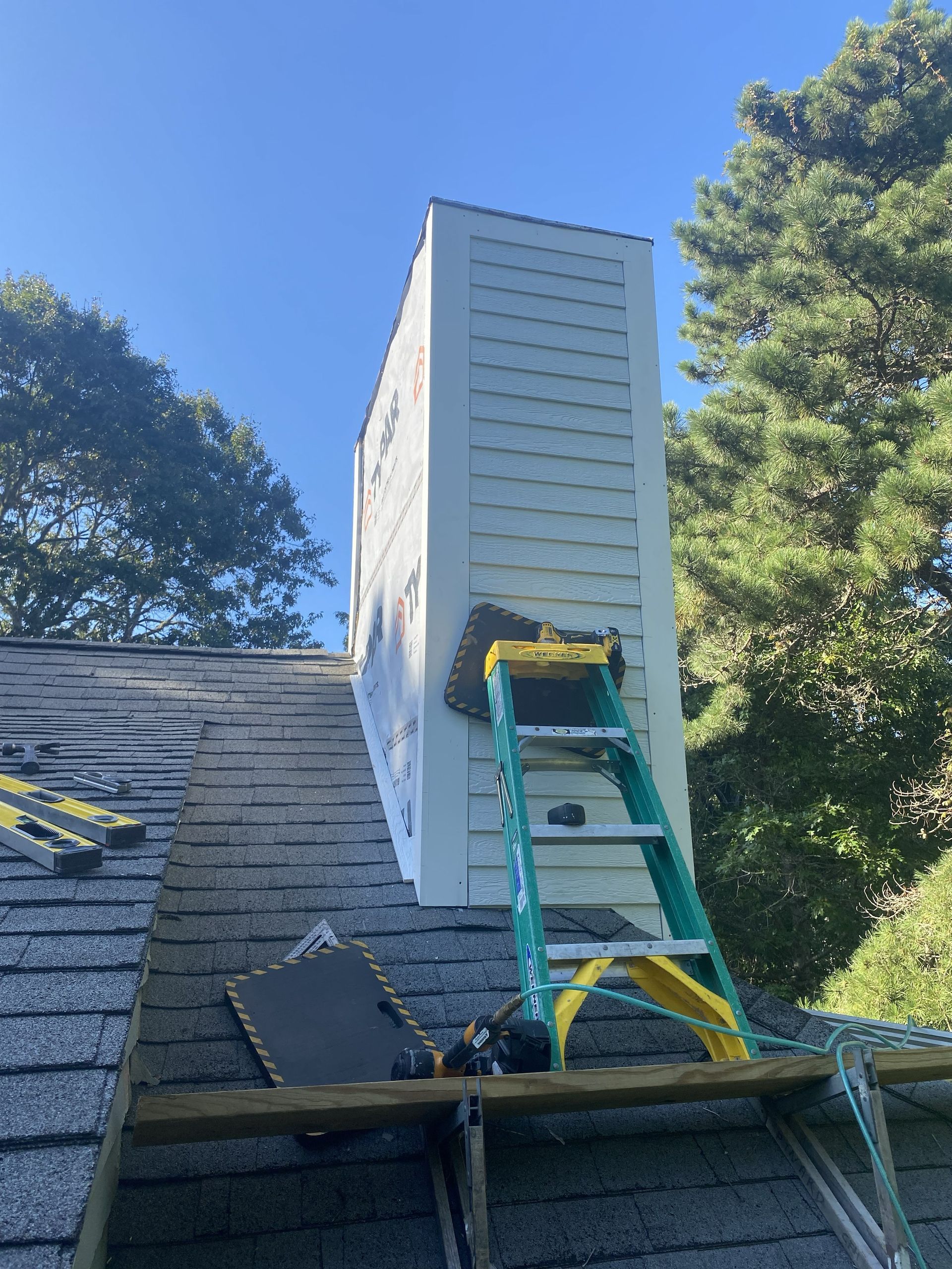 Teal ladder leaning against a rooftop chimney under a blue sky, with trees in the background