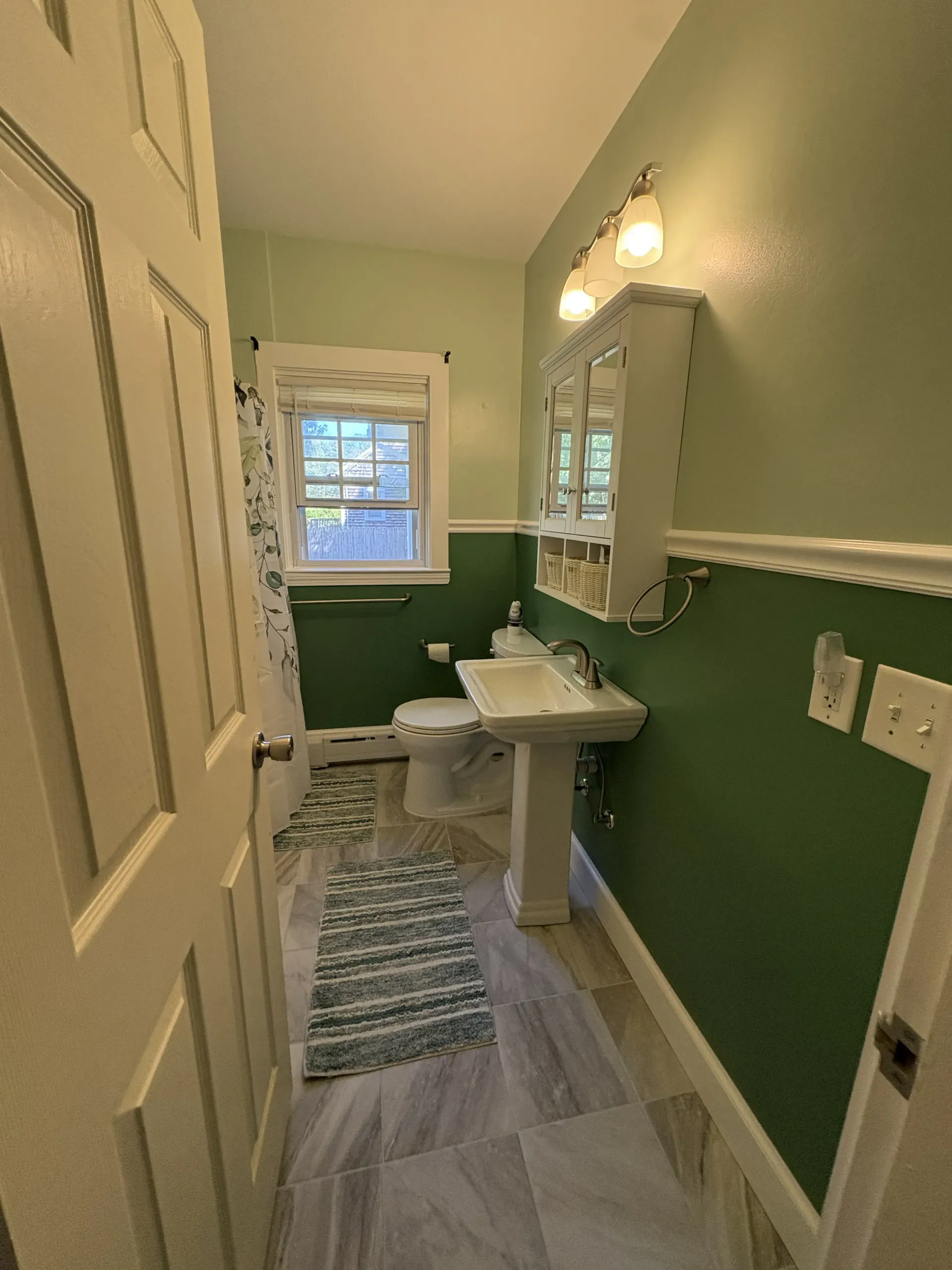 Narrow bathroom with green walls, pedestal sink, mirror, window, and patterned runner rug.