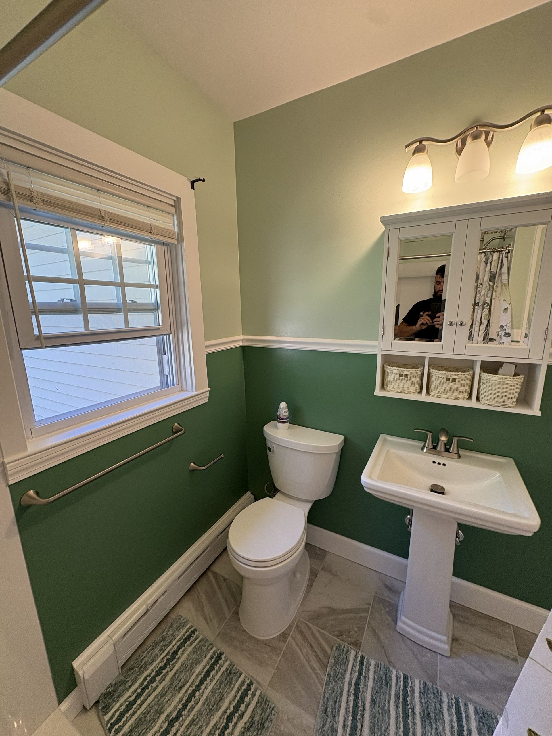 Small bathroom with green walls, white pedestal sink and toilet, window, mirror cabinet, and striped rug.