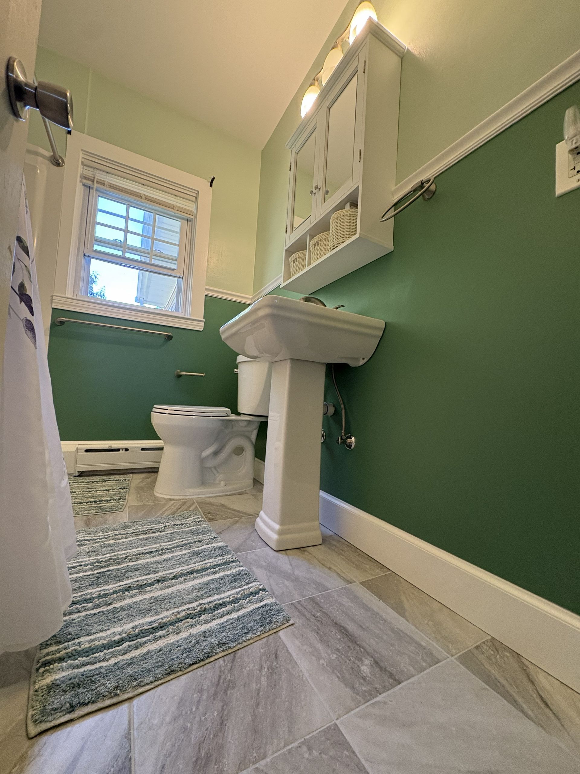 Small bathroom with green walls, pedestal sink, toilet, window, and striped rug