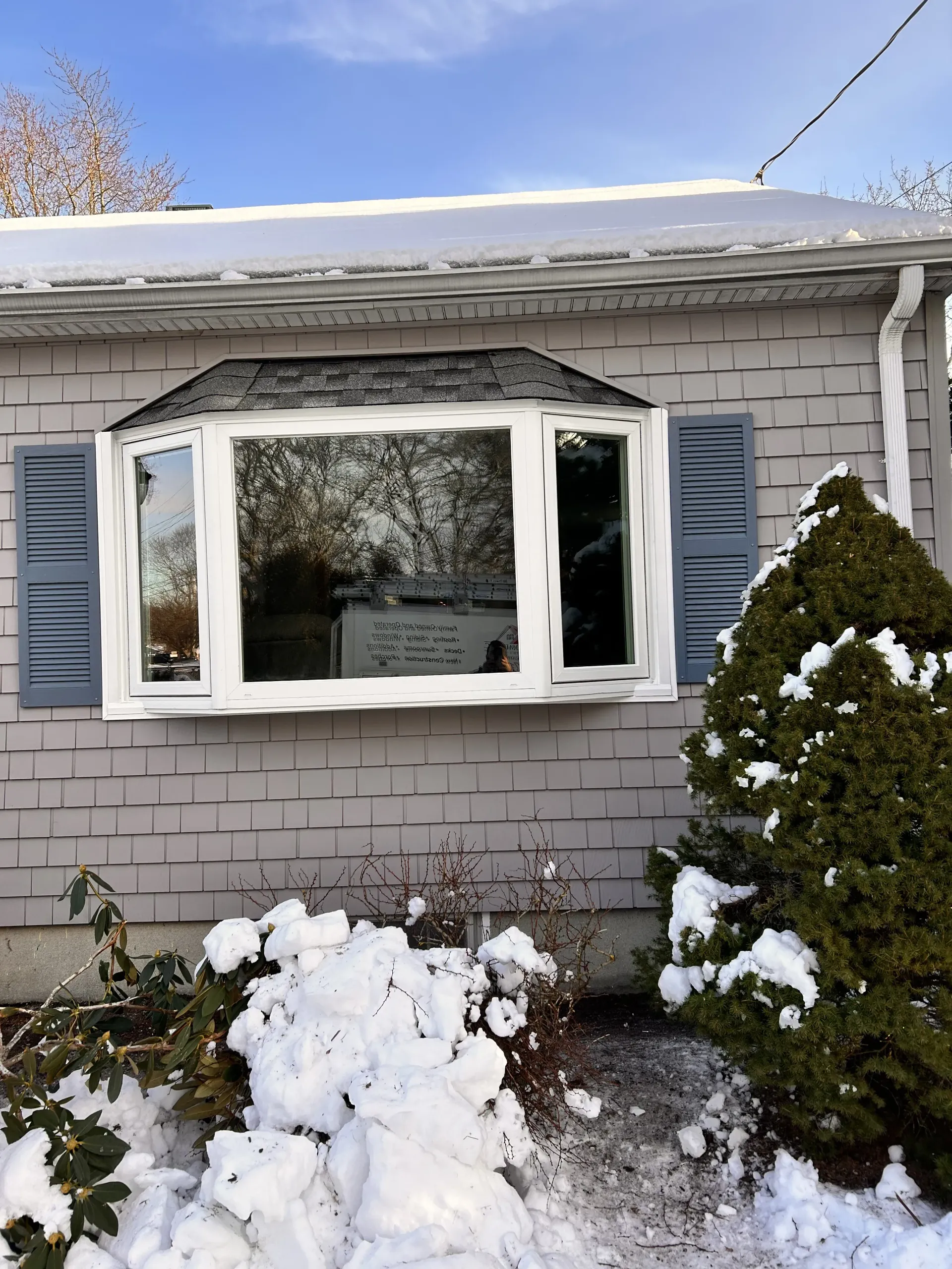 Snow-covered house exterior with a large white-trimmed bay window and shrubs in front