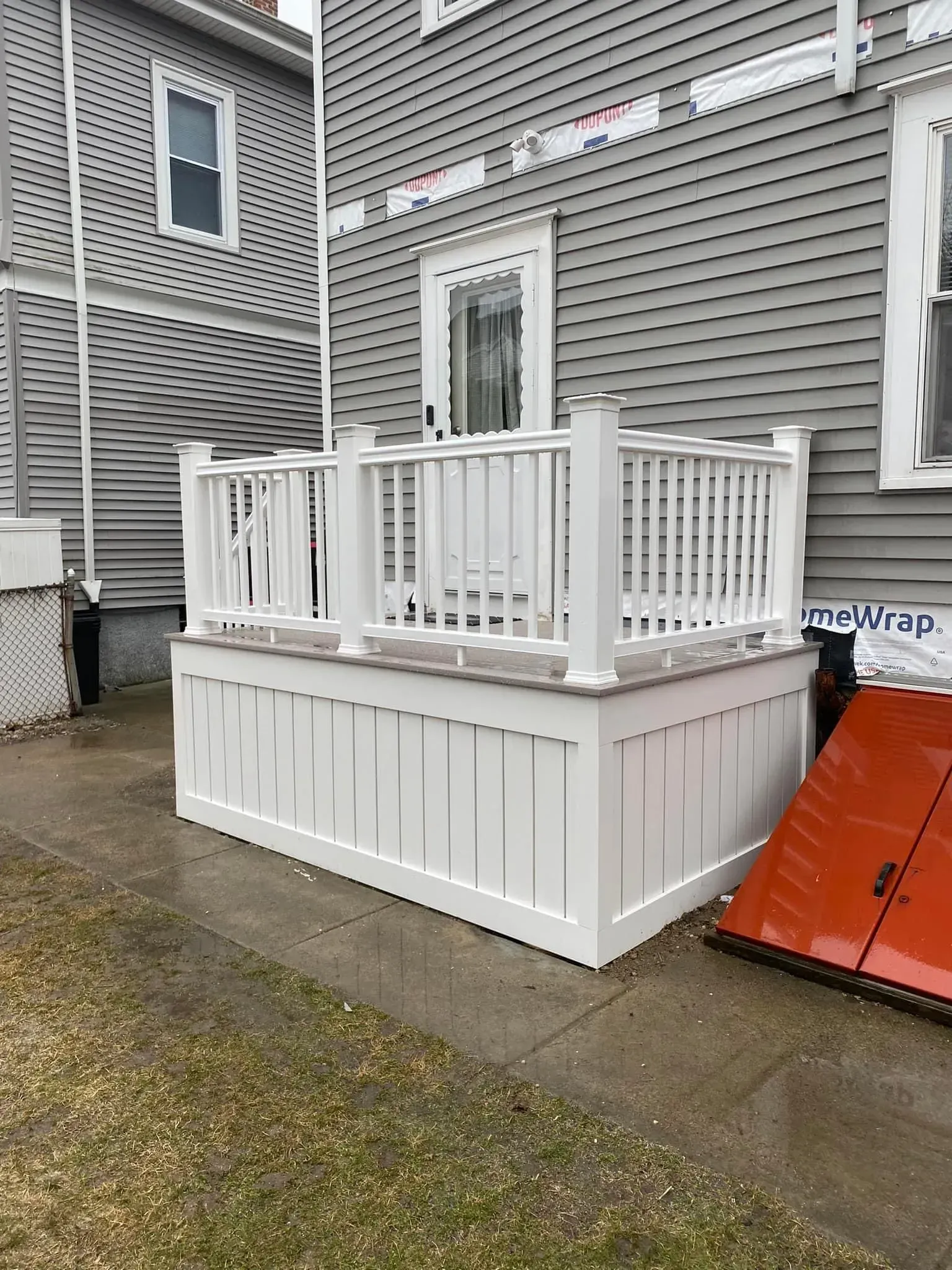 White-railed raised deck attached to a gray house, beside an orange ramp on a wet walkway