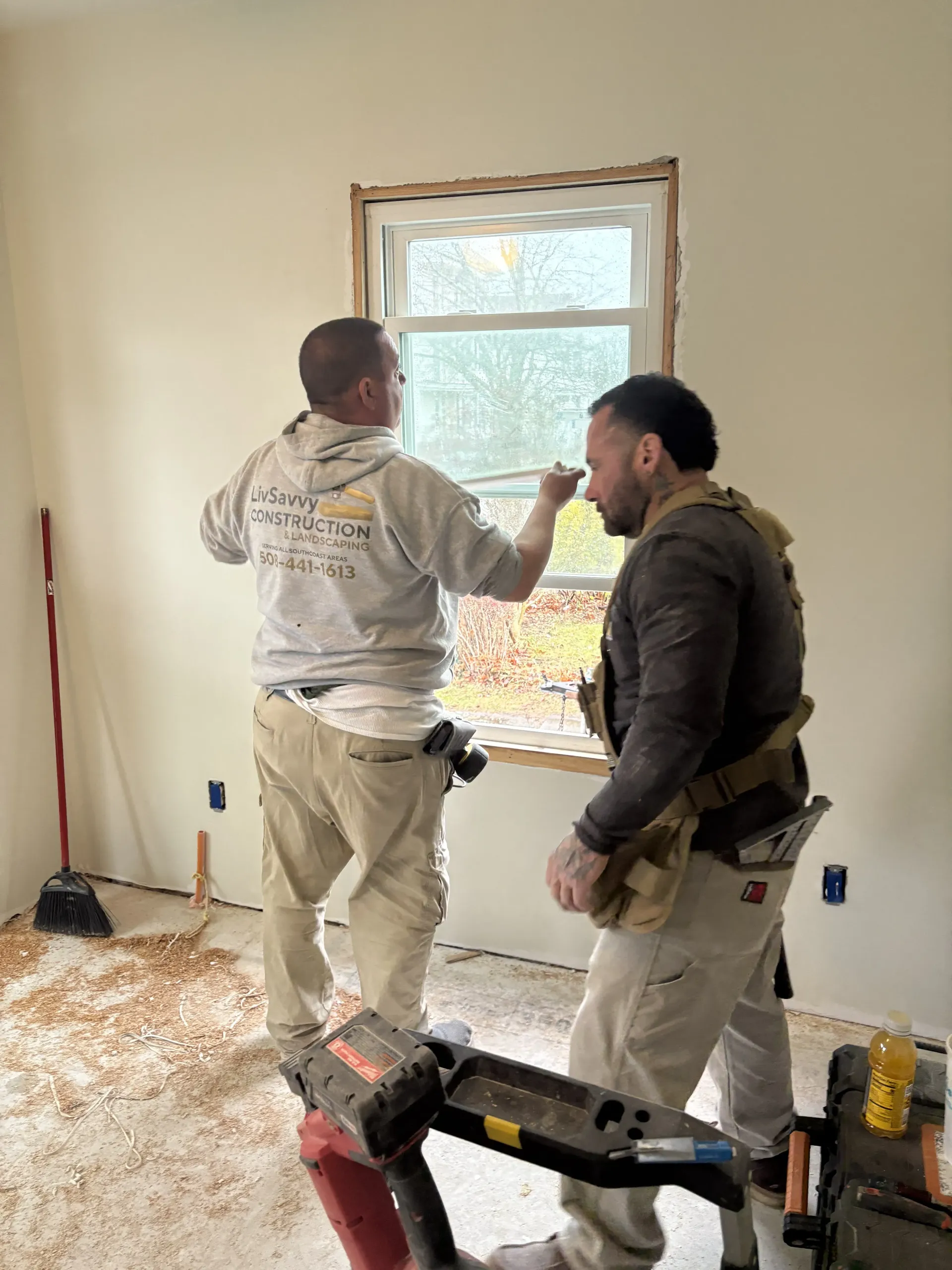 Two workers installing a window frame in a room under renovation, with tools and debris on the floor.
