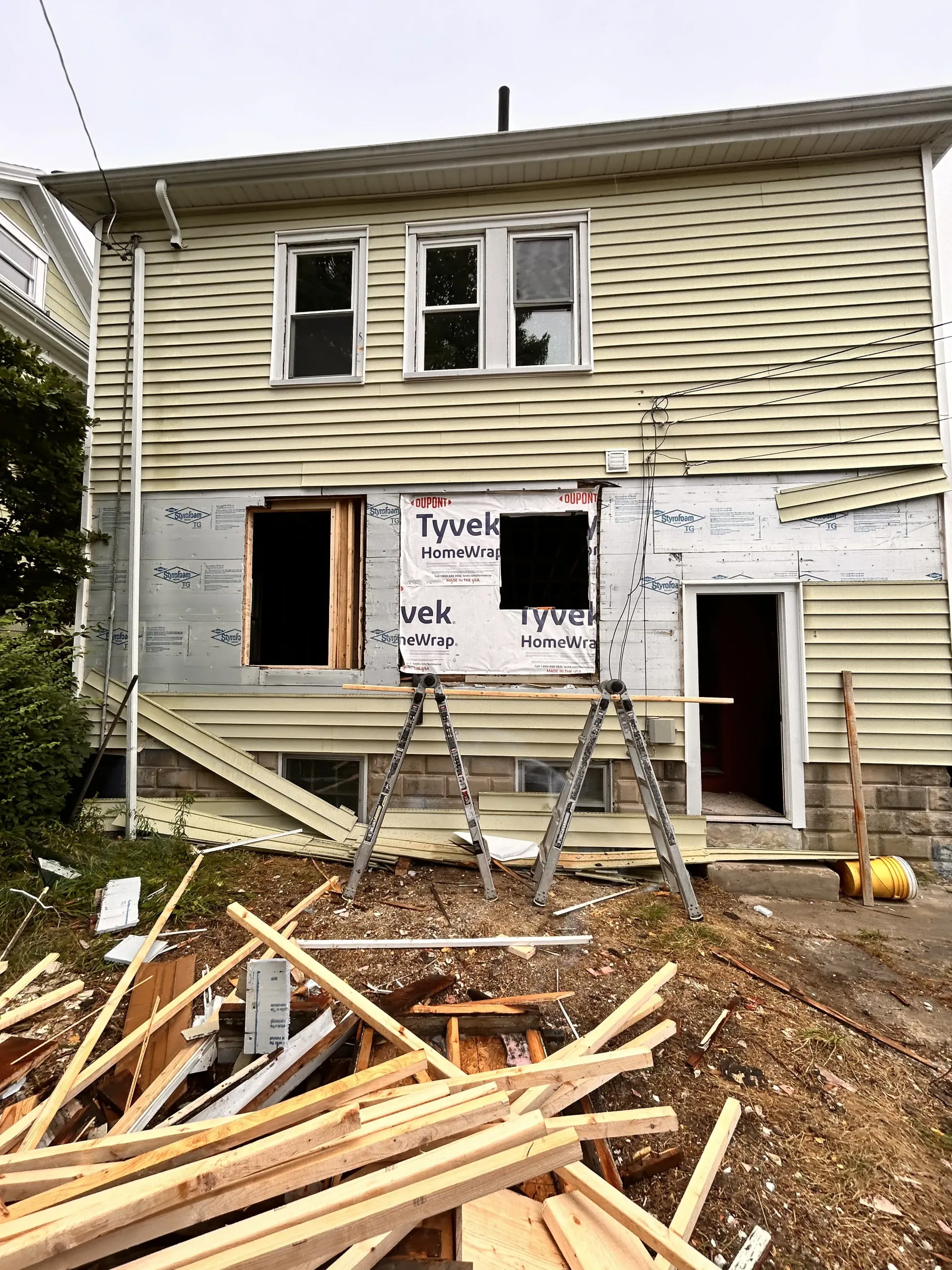 Partially renovated two-story house with open windows, ladders, and wood debris in front yard