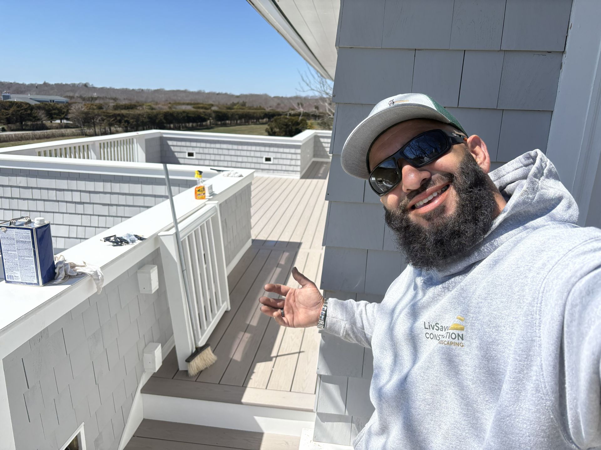 Smiling man in gray hoodie and cap gestures on a rooftop with white walls and sunny hills behind him
