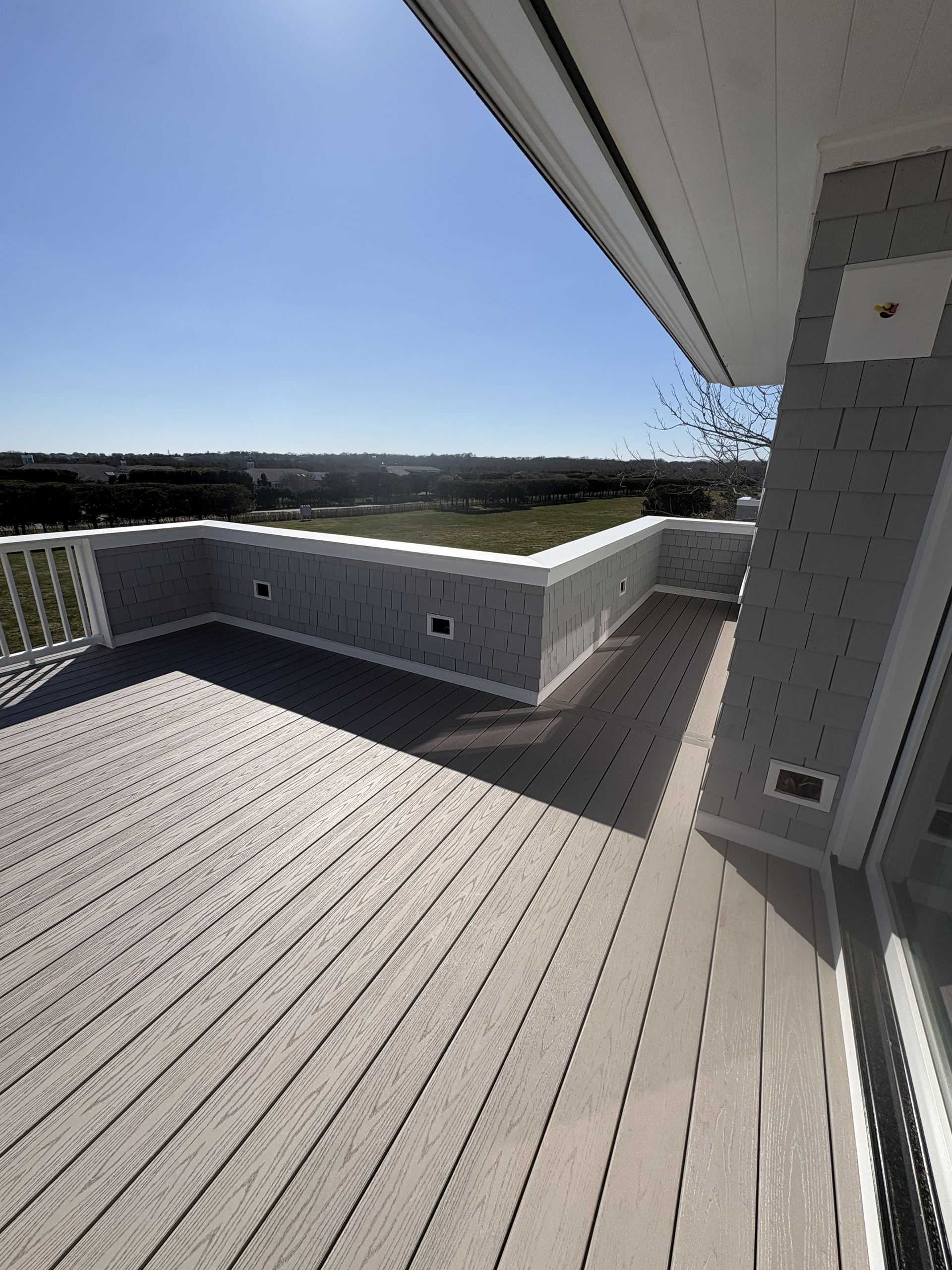 Sunlit rooftop deck with white railing, gray flooring, and a view of fields under a clear blue sky