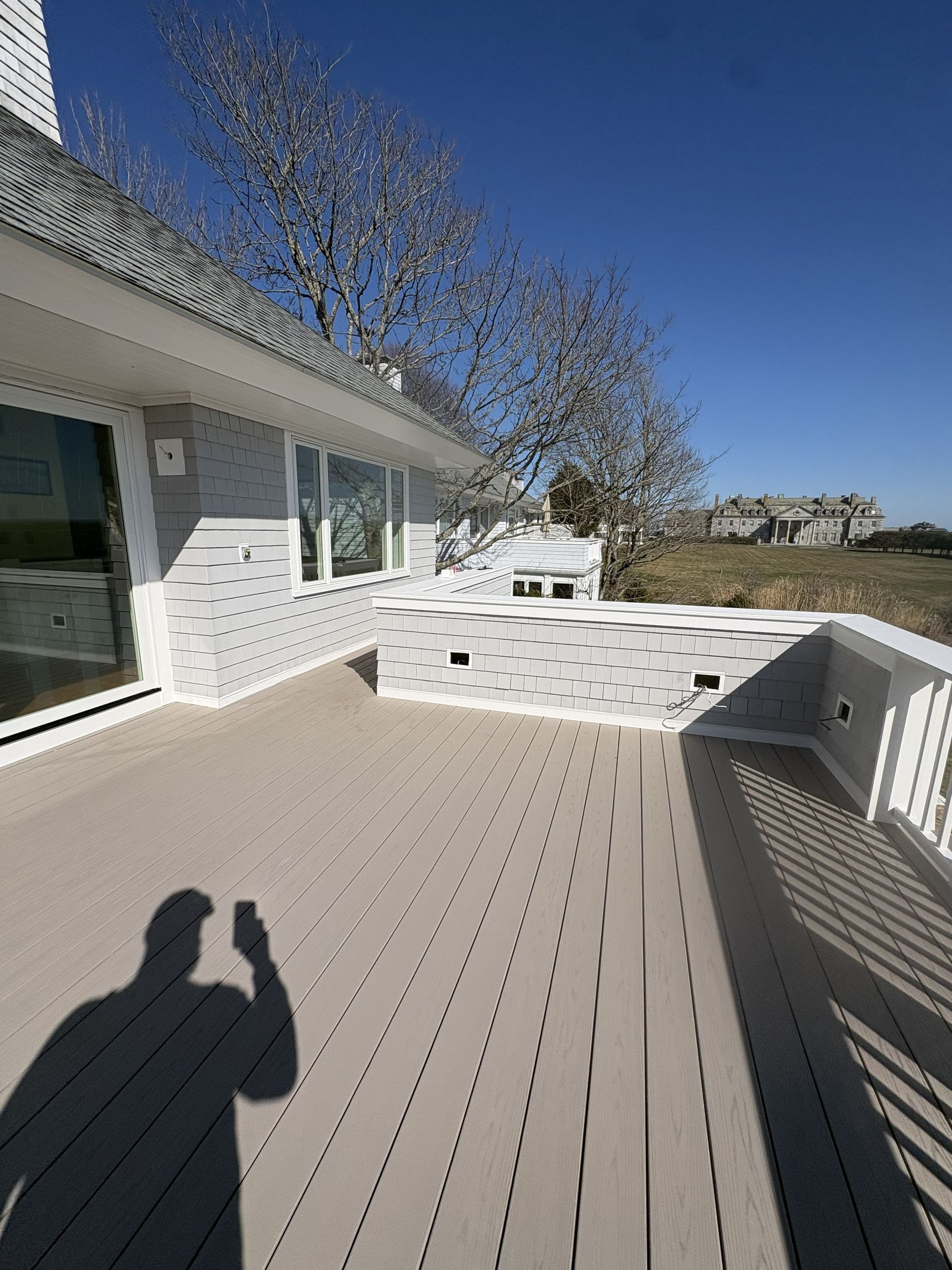 Sunny rooftop deck with white railing, gray floorboards, and a house wall with windows and trees beyond