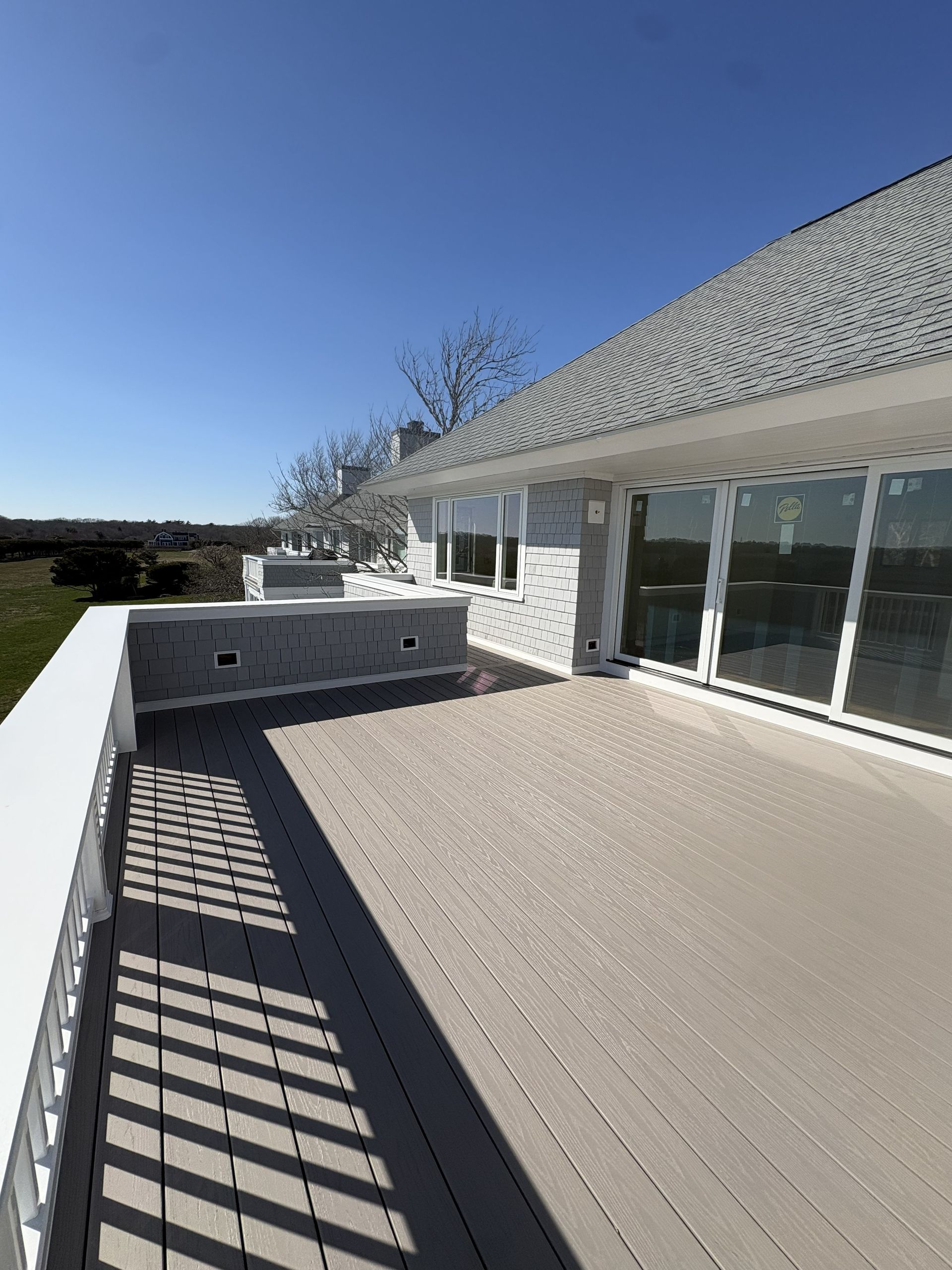 Sunny rooftop deck with gray pavers, glass doors, and railing shadows beside a white house
