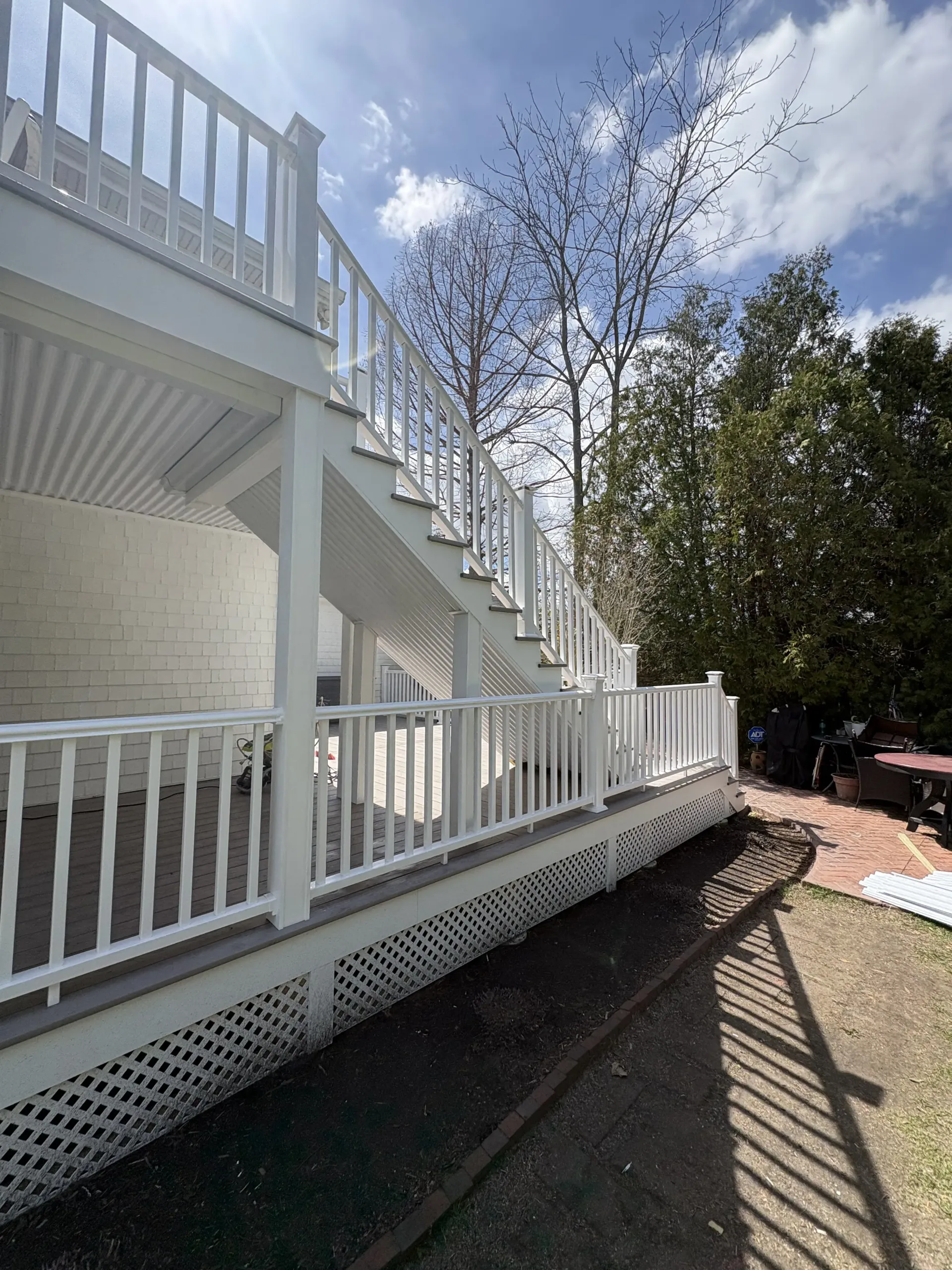 White porch and staircase with railings along a sunny walkway beside a stone building and trees