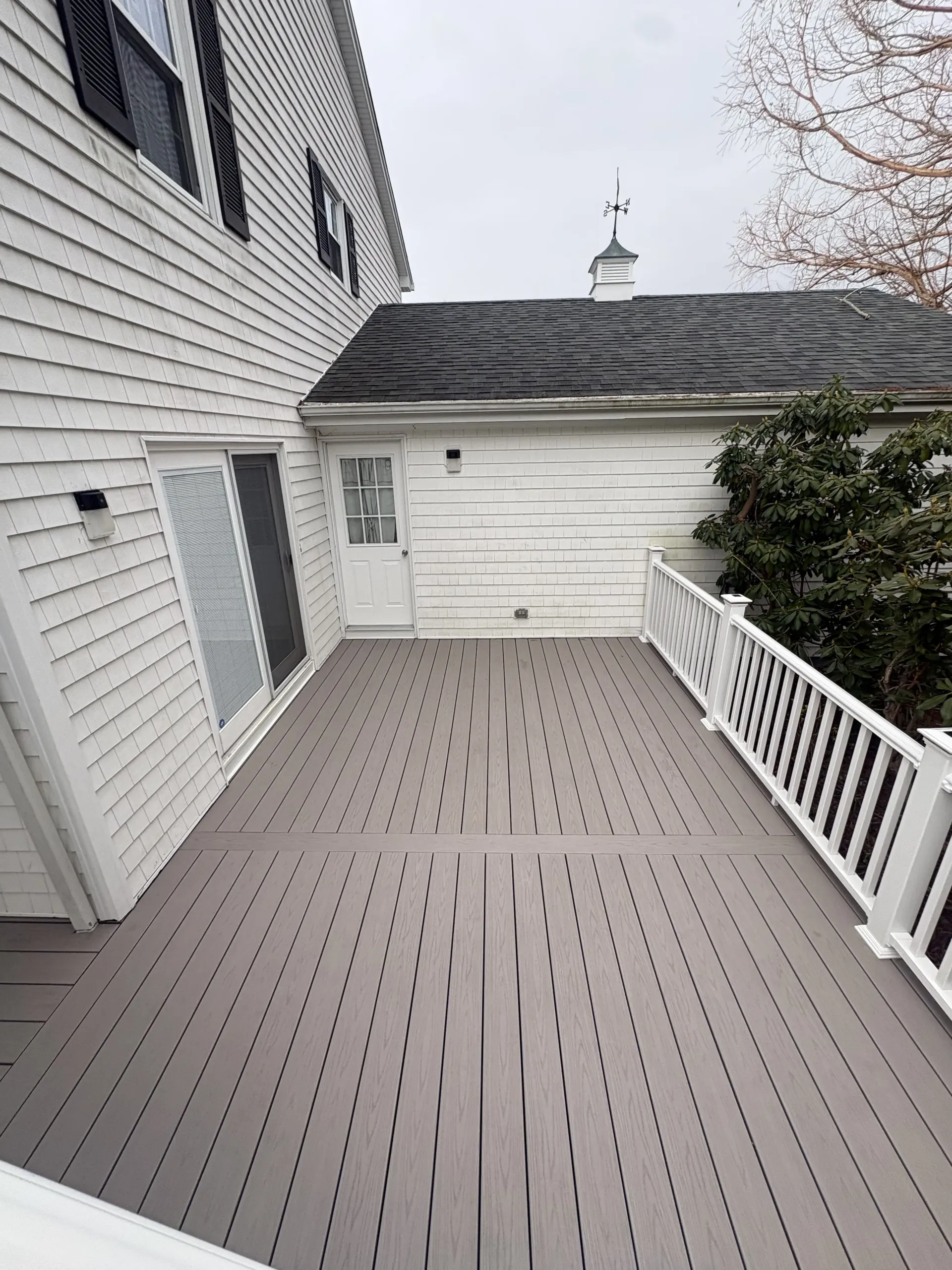 Narrow gray composite deck beside a white house, with railing and small shed-like structure at the end.