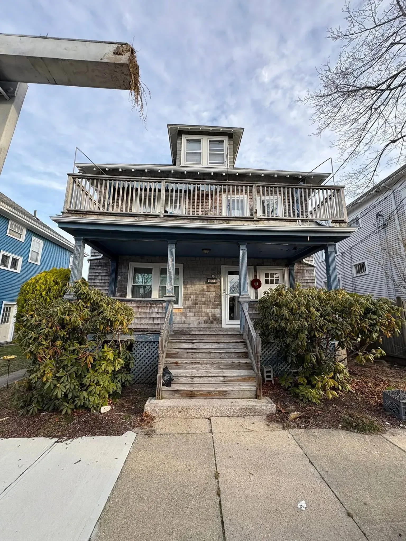 Front view of a weathered two-story house with a front porch, steps, and bushes along a sidewalk.