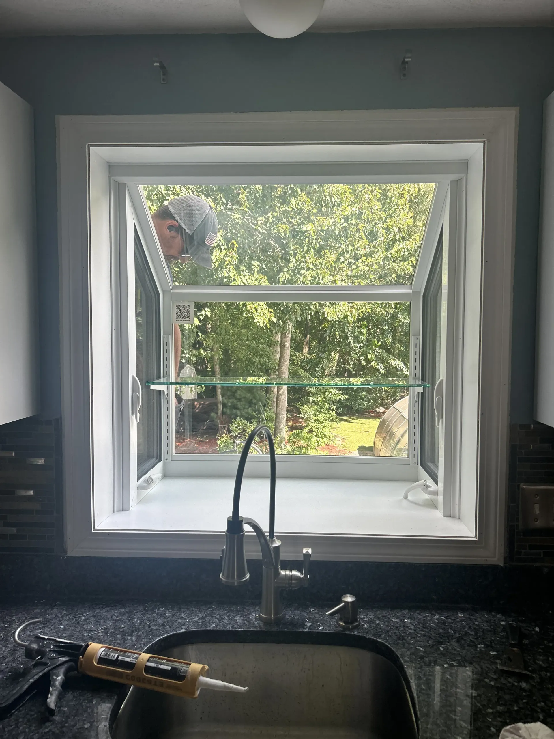 Kitchen sink with black countertop looking out through a large window to trees and a person outside