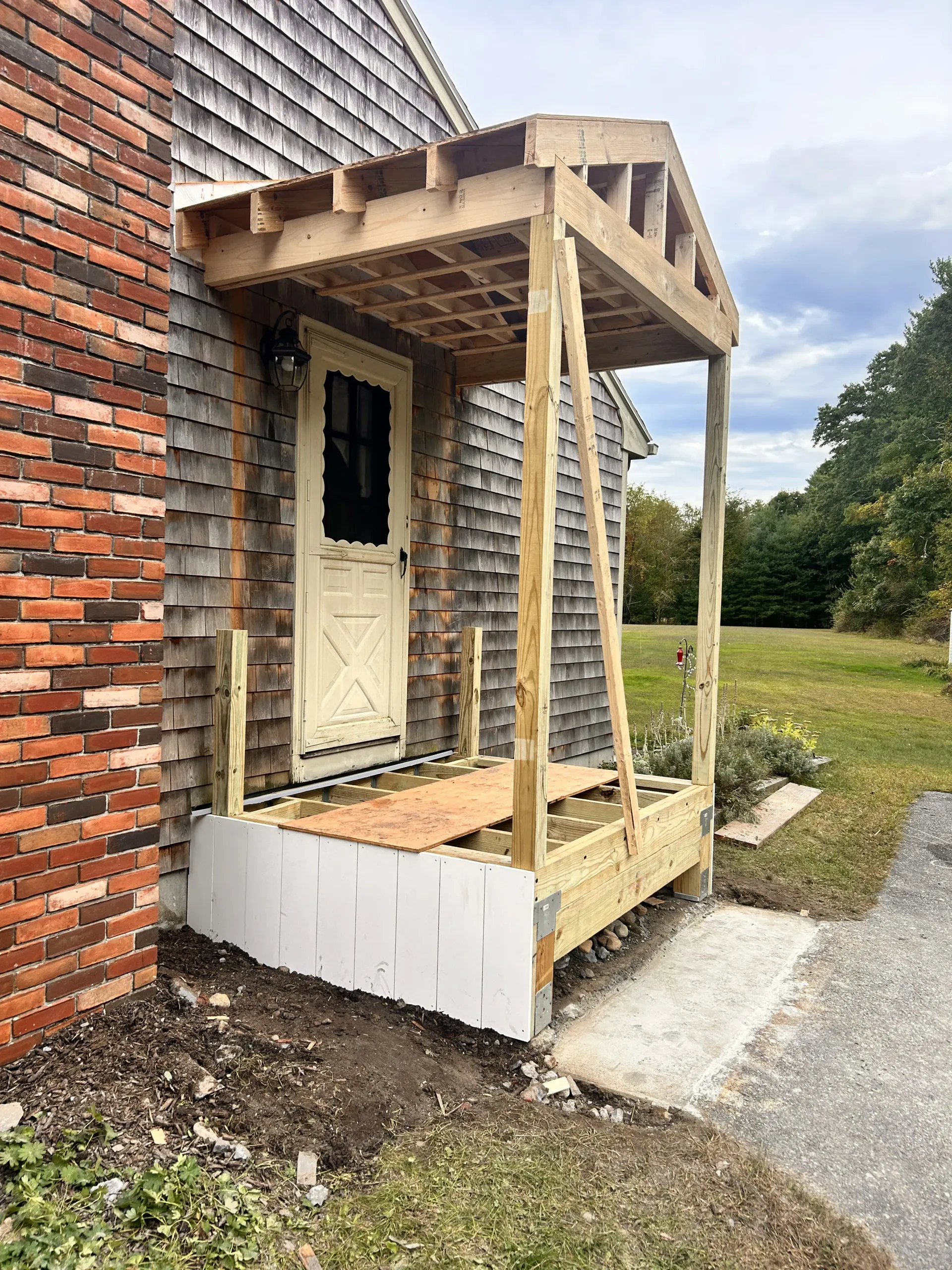 Small brick house entry with a rough wooden porch and steps beside a gravel path.