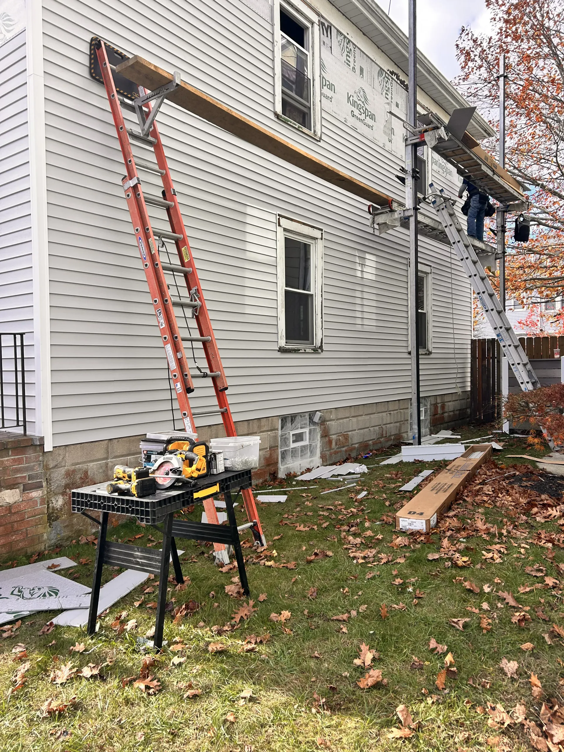 Two ladders against a house with a worker on the roof and leaves scattered on the lawn