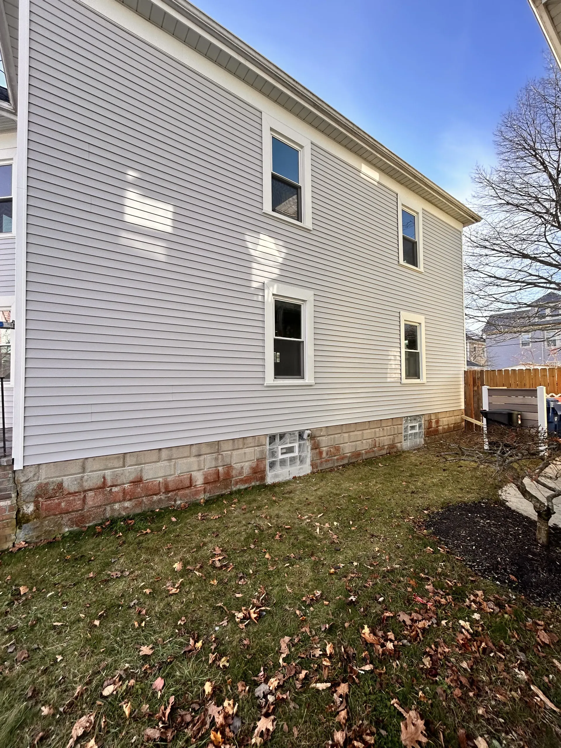 White vinyl-sided house exterior with two windows and a small basement window above a leaf-covered yard
