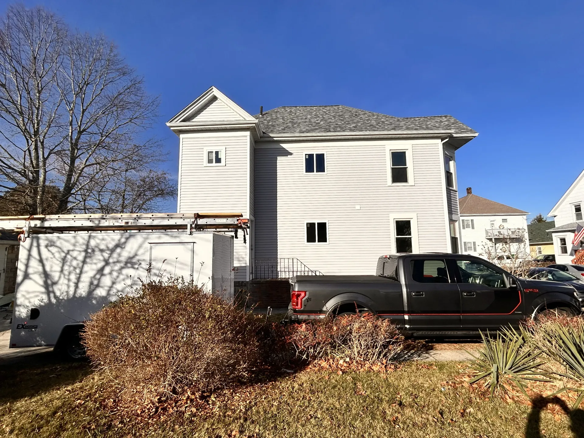 White two-story house with a black pickup truck, garage, and piles of leaves in the yard under a blue sky