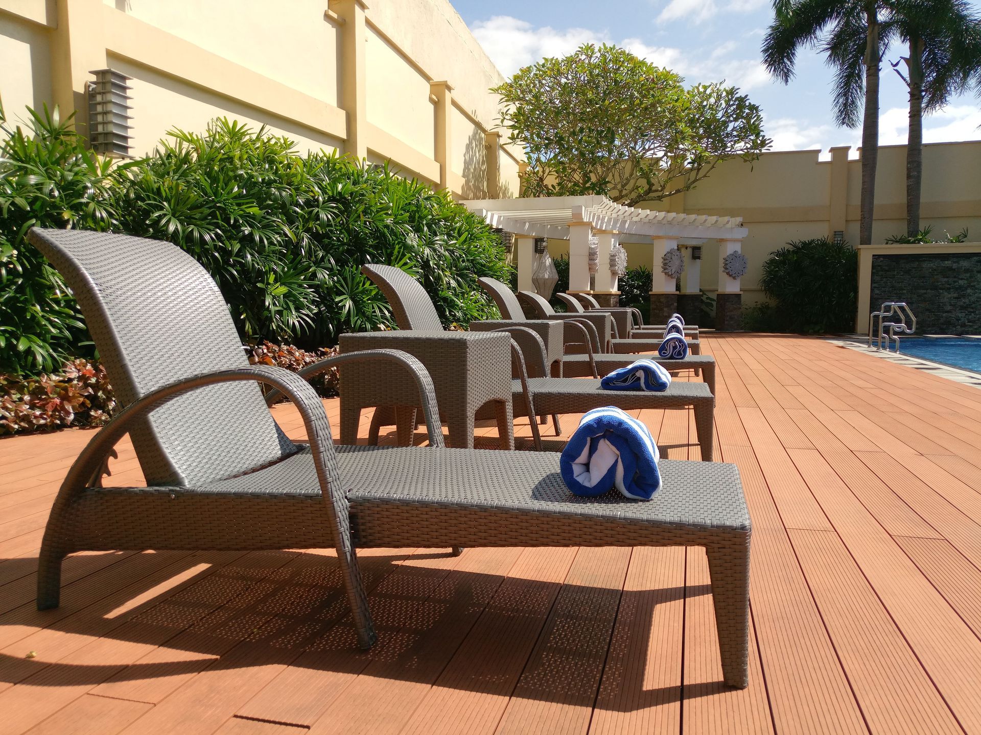 Lounge chairs by a sunny pool deck with a blue towel on the nearest chair, surrounded by greenery and buildings