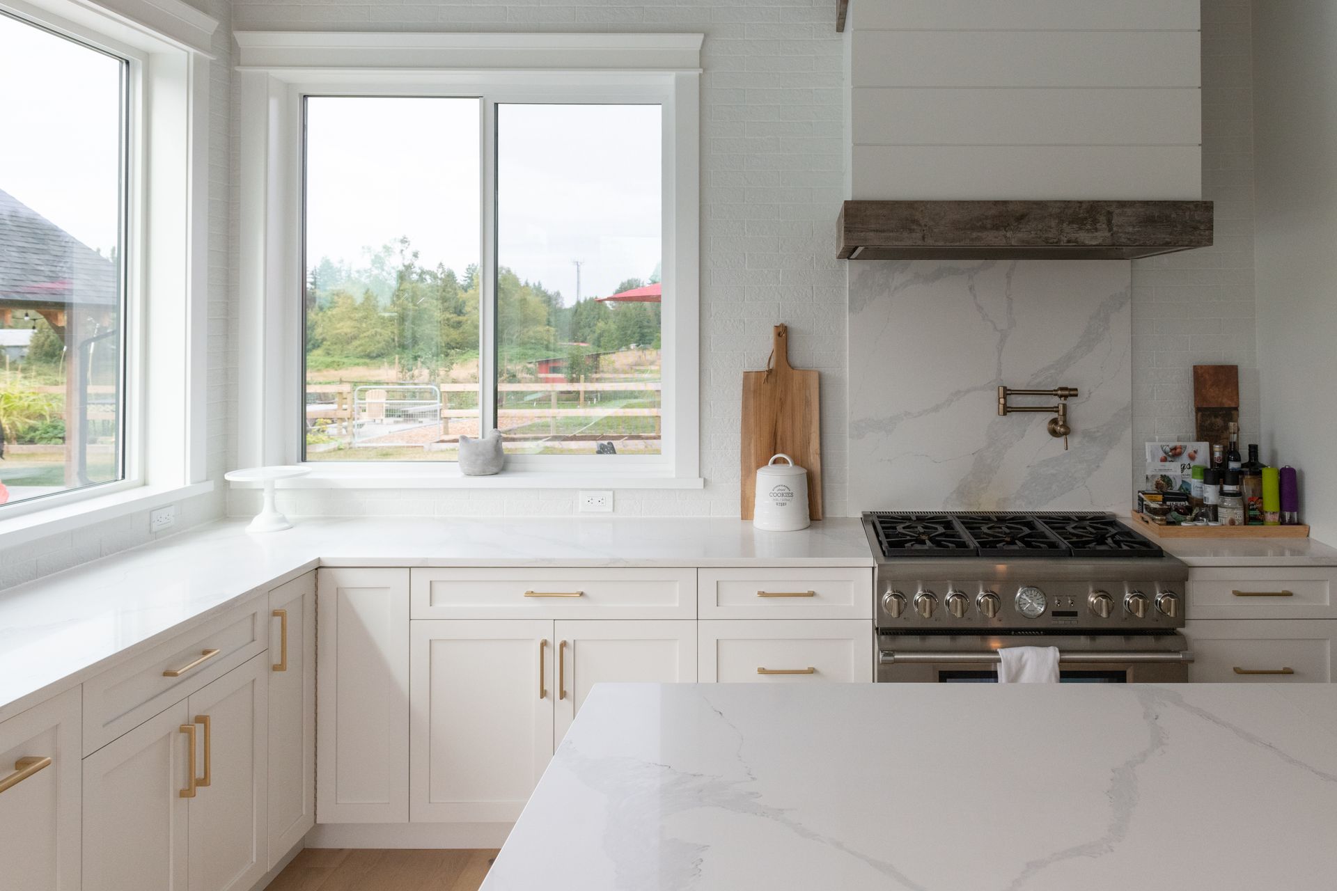 Bright white kitchen with marble island, large windows, and stainless steel stove under a wooden range hood