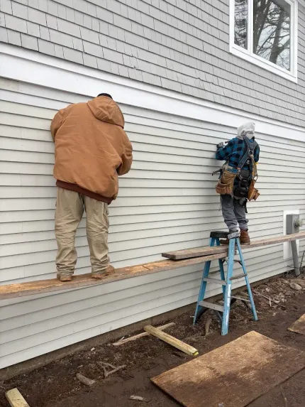 Two workers installing horizontal siding on a house, one on a plank and one on a ladder.