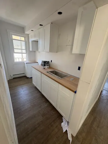Narrow white galley kitchen with wood countertop, sink, cabinets, and window at the end