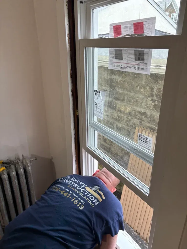 Worker leaning out an open window during construction, with tools and a radiator visible inside.