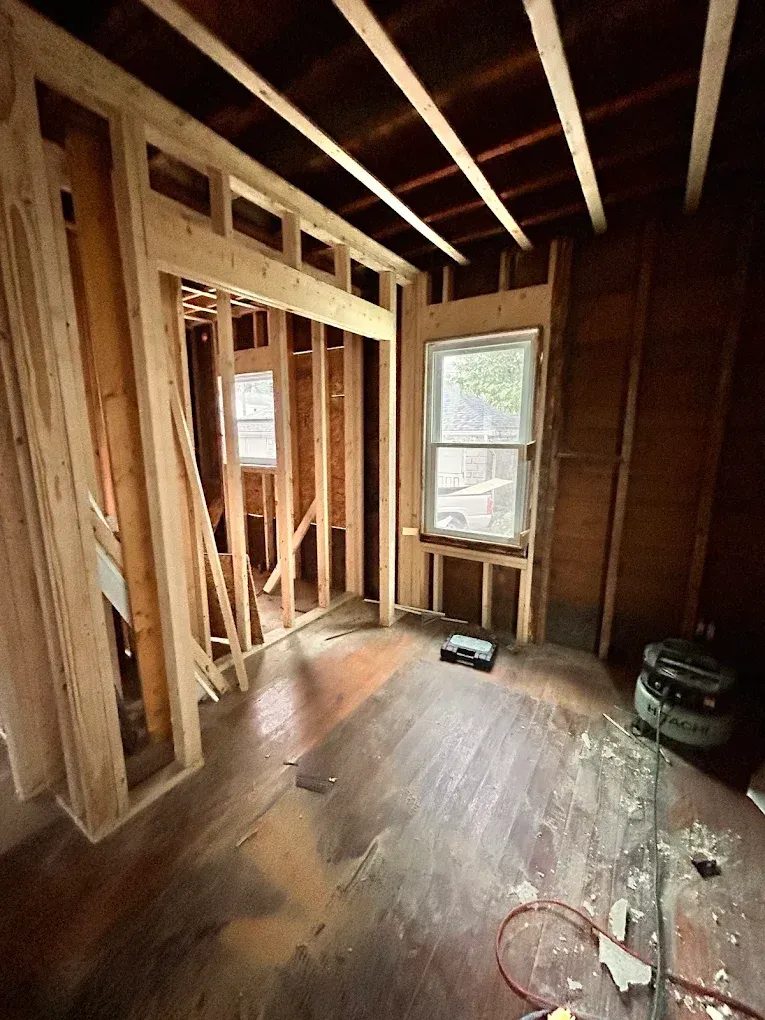 Unfinished room under construction with exposed studs, beams, and two windows on a dusty floor.