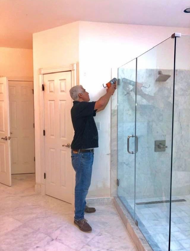 Man installing hardware on a glass shower door in a bathroom with white walls and tiled floor