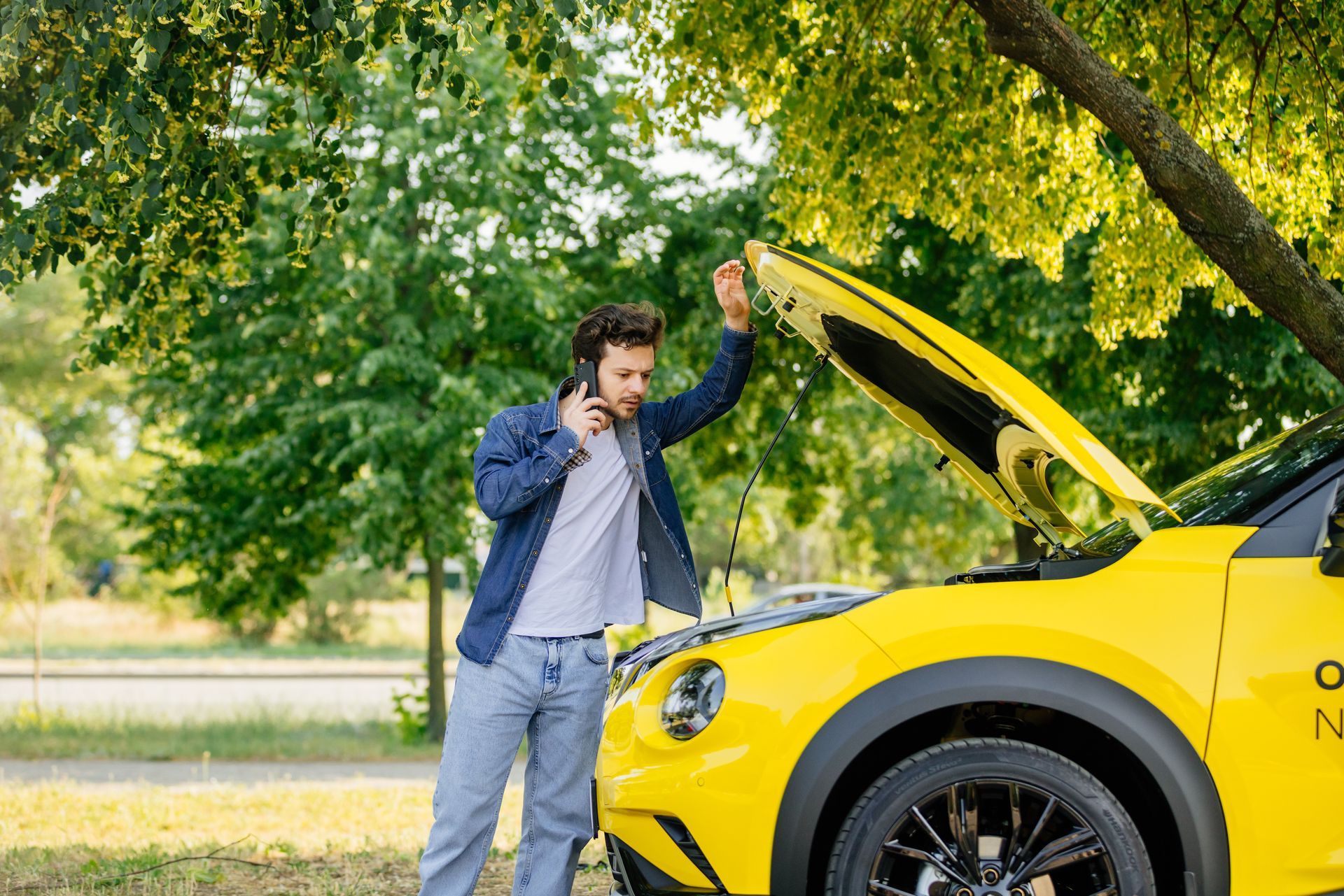 Man checking under the hood of a yellow car in a leafy outdoor setting